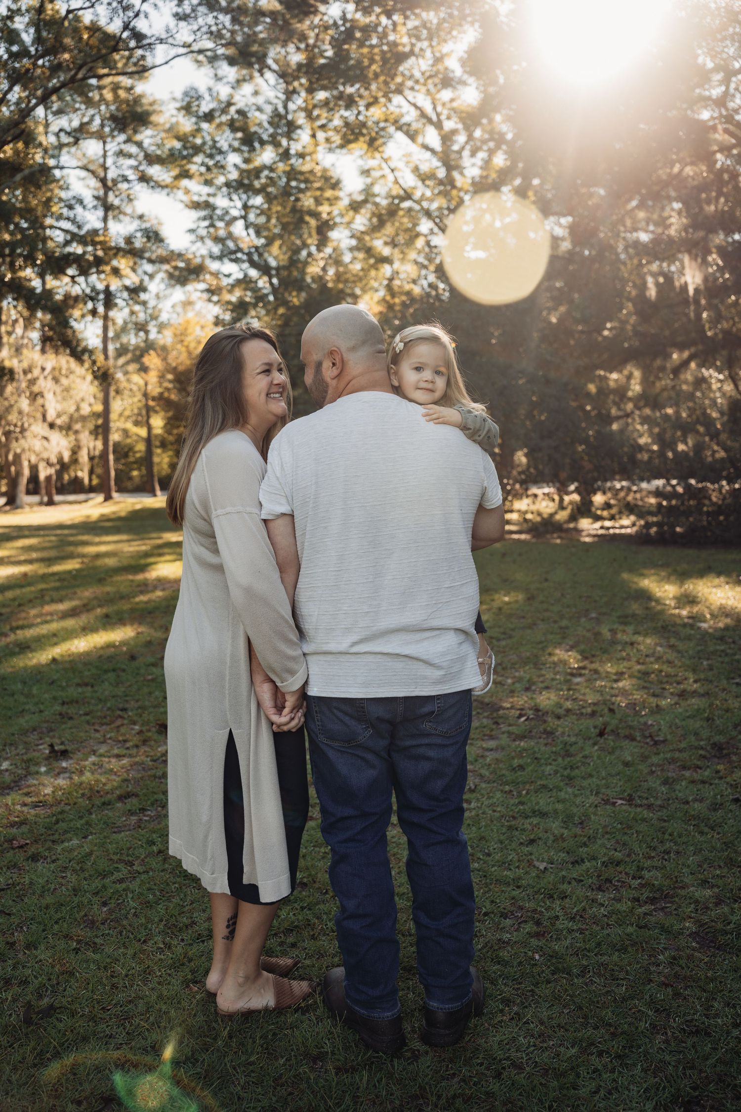 A family of three shares a tender moment outdoors, with golden sunlight filtering through trees as parents hold their toddler.