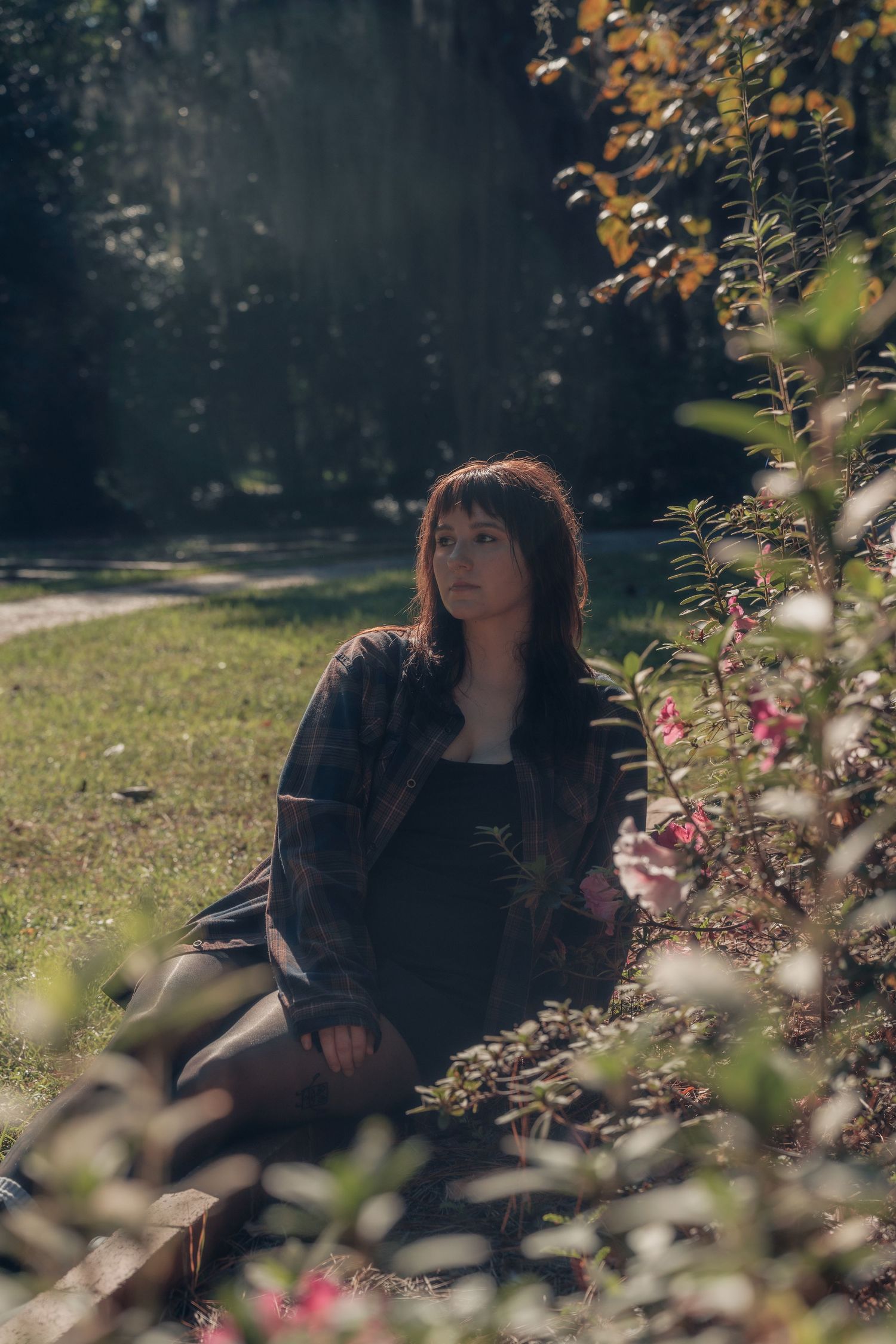 Young woman with auburn hair sitting peacefully beside flowering bushes in a sunlit park, wearing a dark floral jacket.