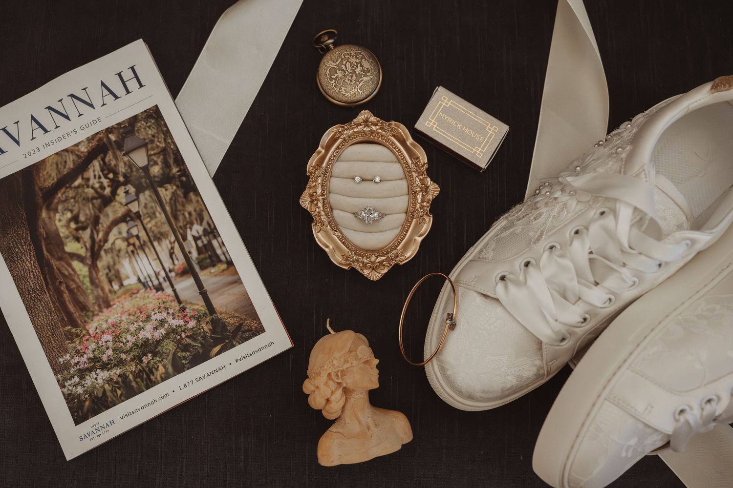 Flat lay of wedding rings in gold frame, white sneakers, Savannah magazine, pocket watch, and cameo brooch on dark wood.