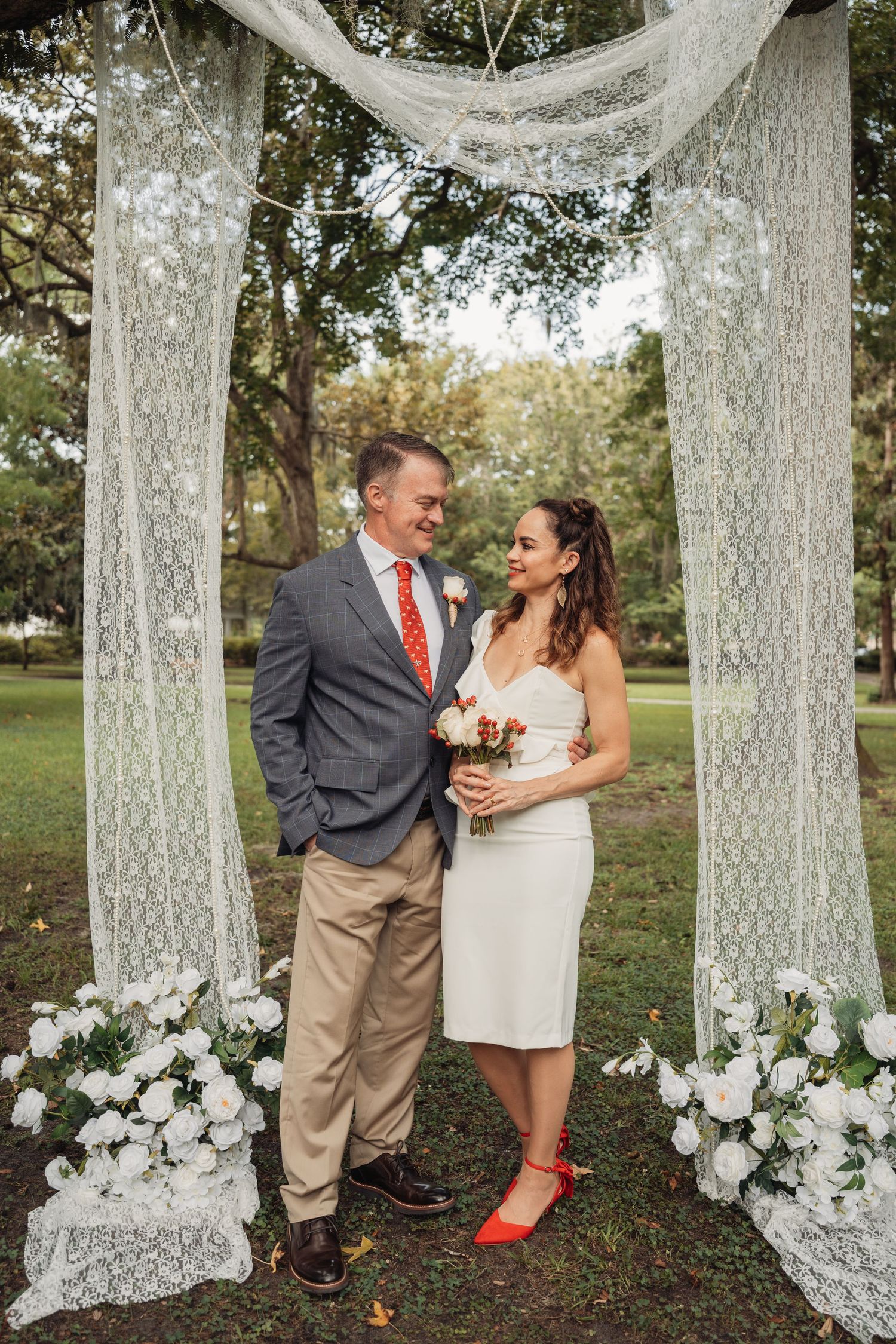 A bride in a white dress with red heels holds flowers beside her groom under a lace wedding arch.