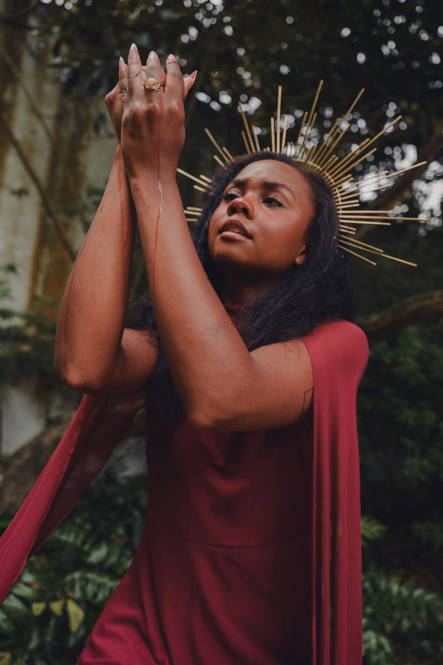 A woman in a red dress wearing a gold halo crown raises her clasped hands upward in a garden setting.