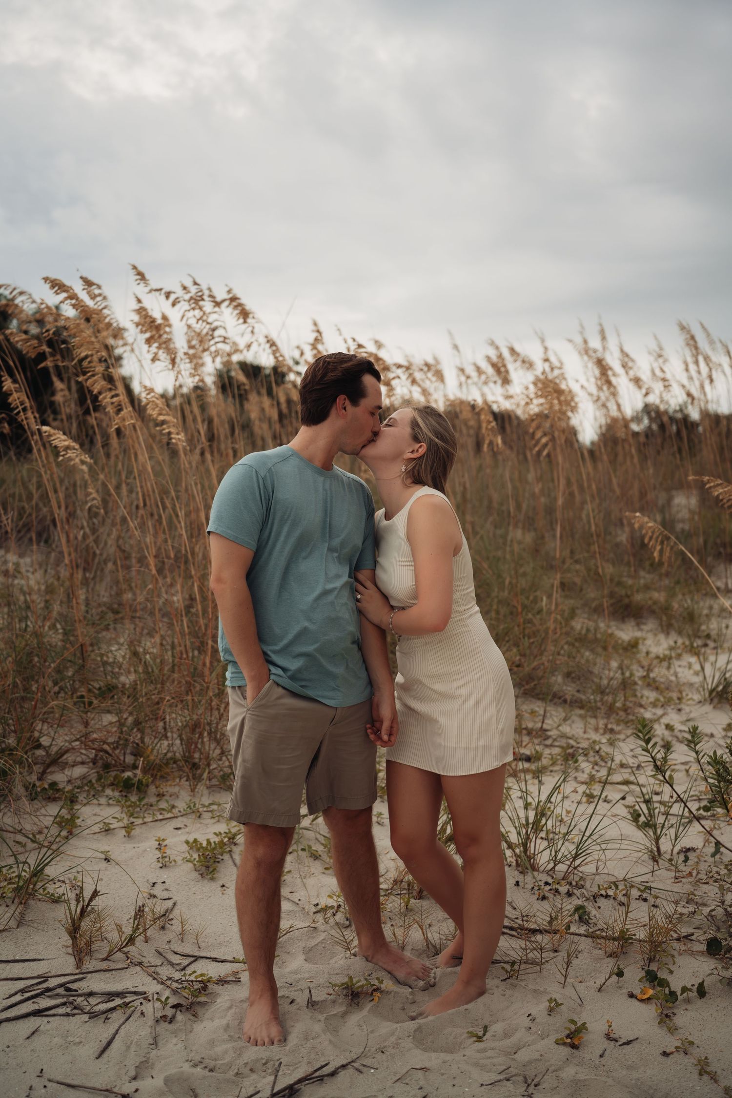 Couple kissing on sandy beach dunes surrounded by sea oats grass under cloudy sky, man in teal shirt and woman in white dress.