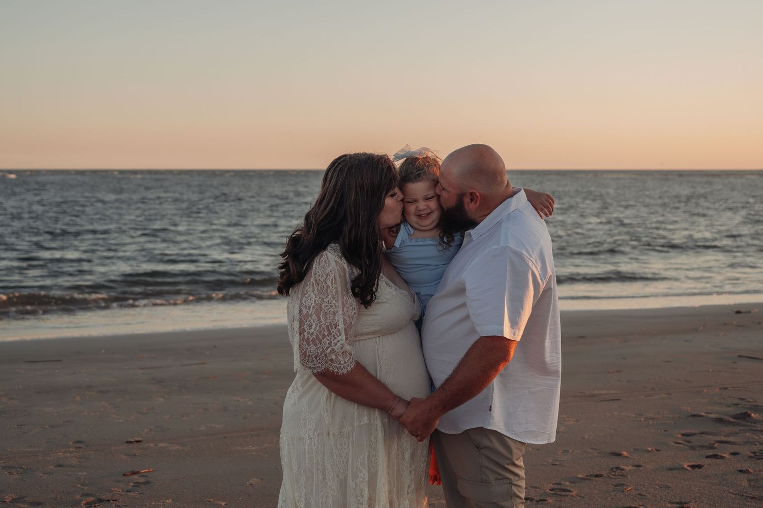 Expectant couple in white embrace their young child on a sandy beach at sunset, with calm ocean waves and a soft pastel sky behind them.
