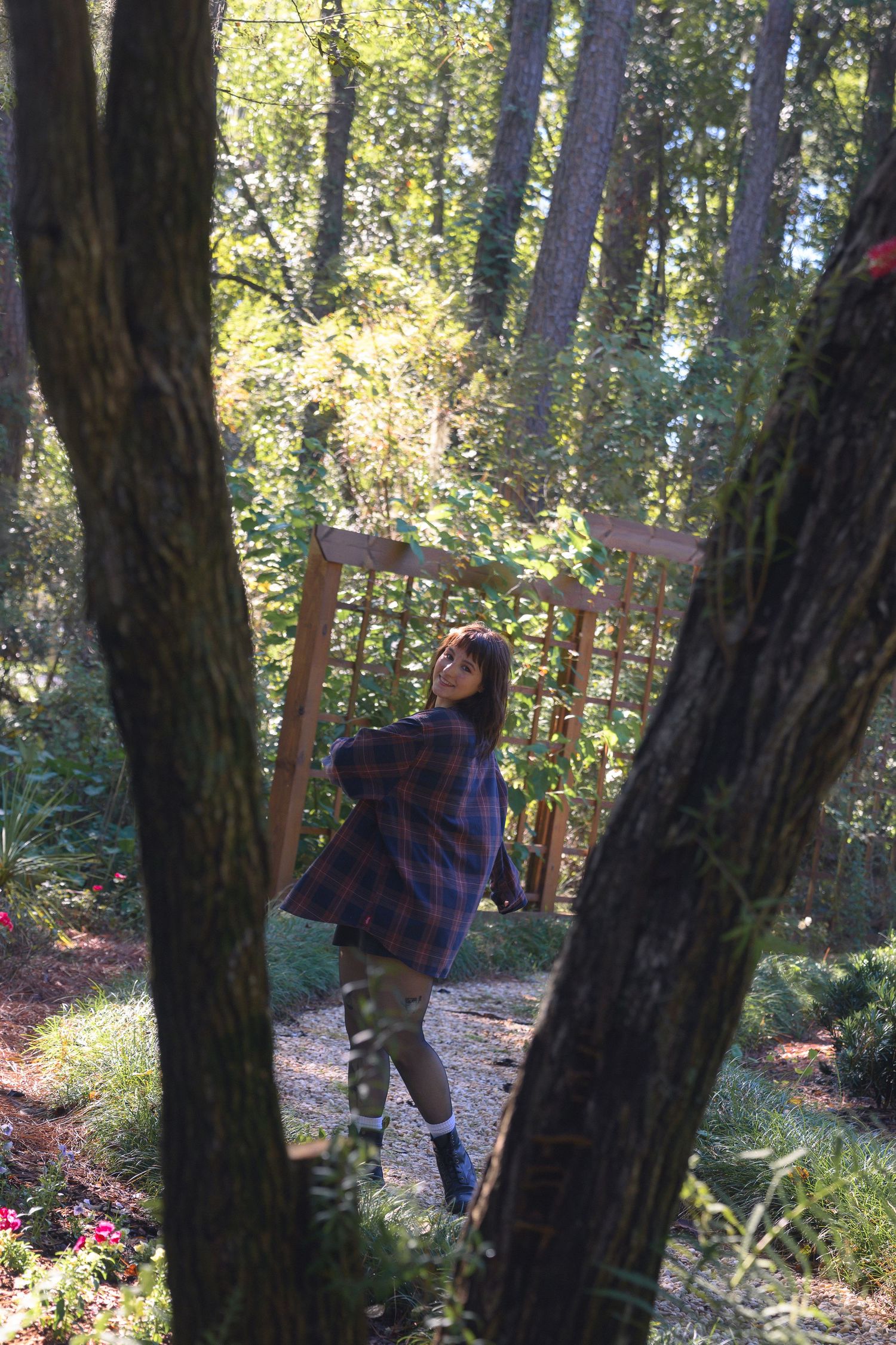 A young woman in a plaid shirt poses among tall trees in a sunlit forest garden with wooden structures.
