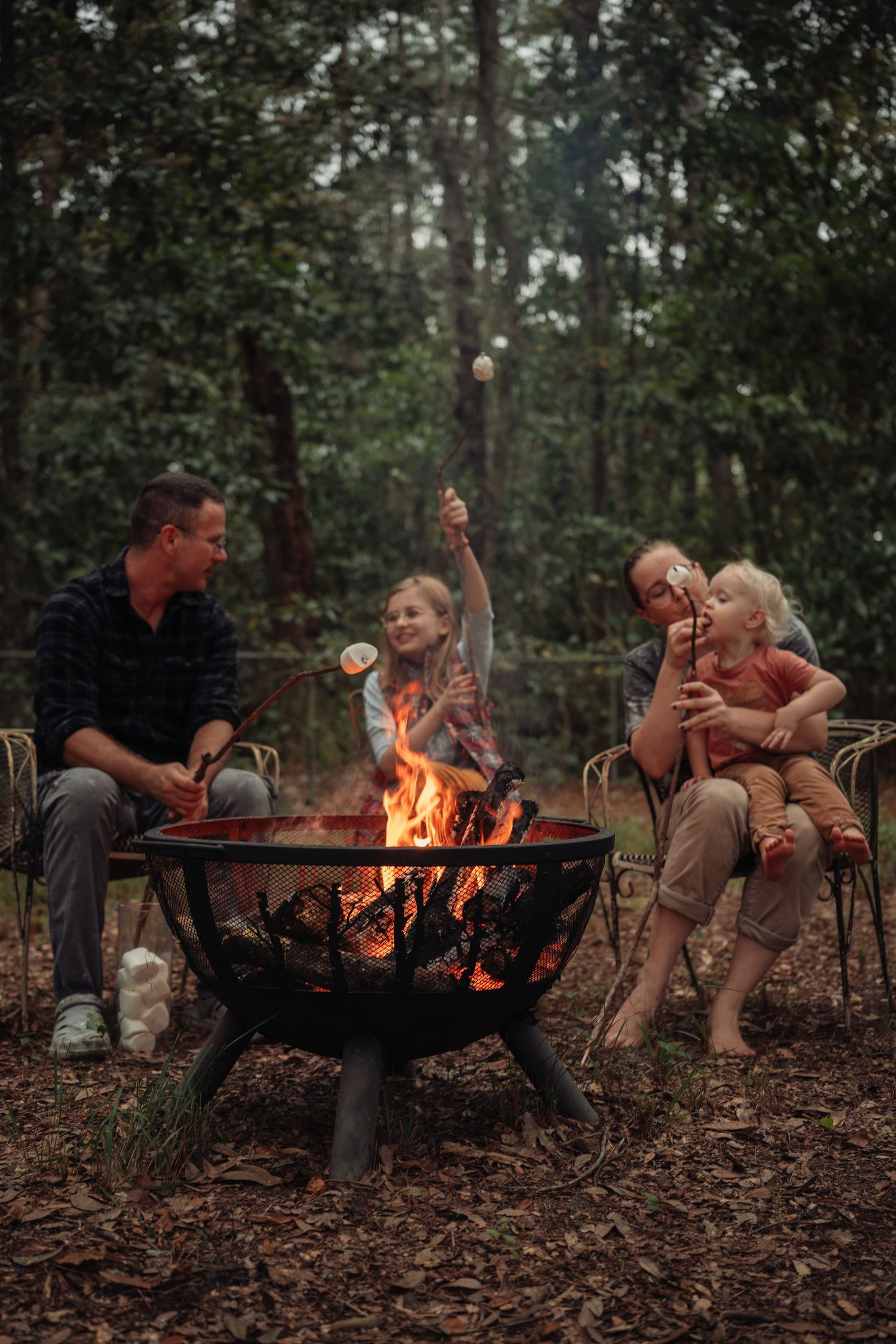 Family roasting marshmallows over a fire pit in a forest setting.