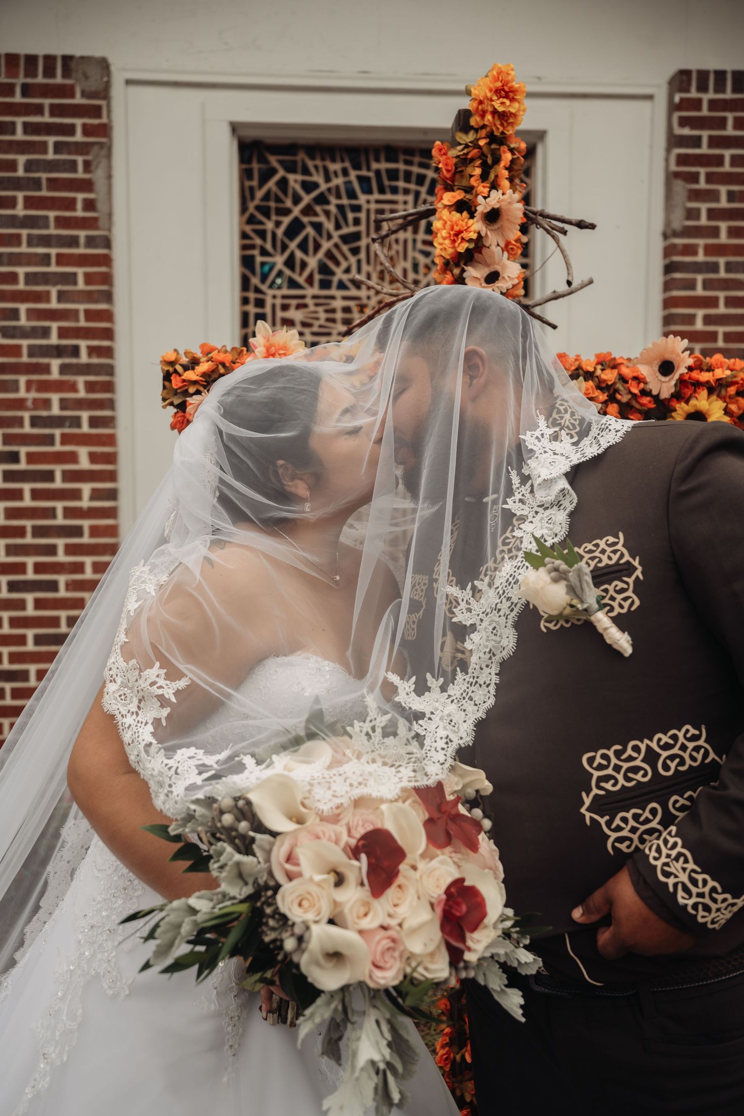 Bride and groom sharing a kiss under a lace veil, holding a bouquet of white and red roses at a fall wedding.