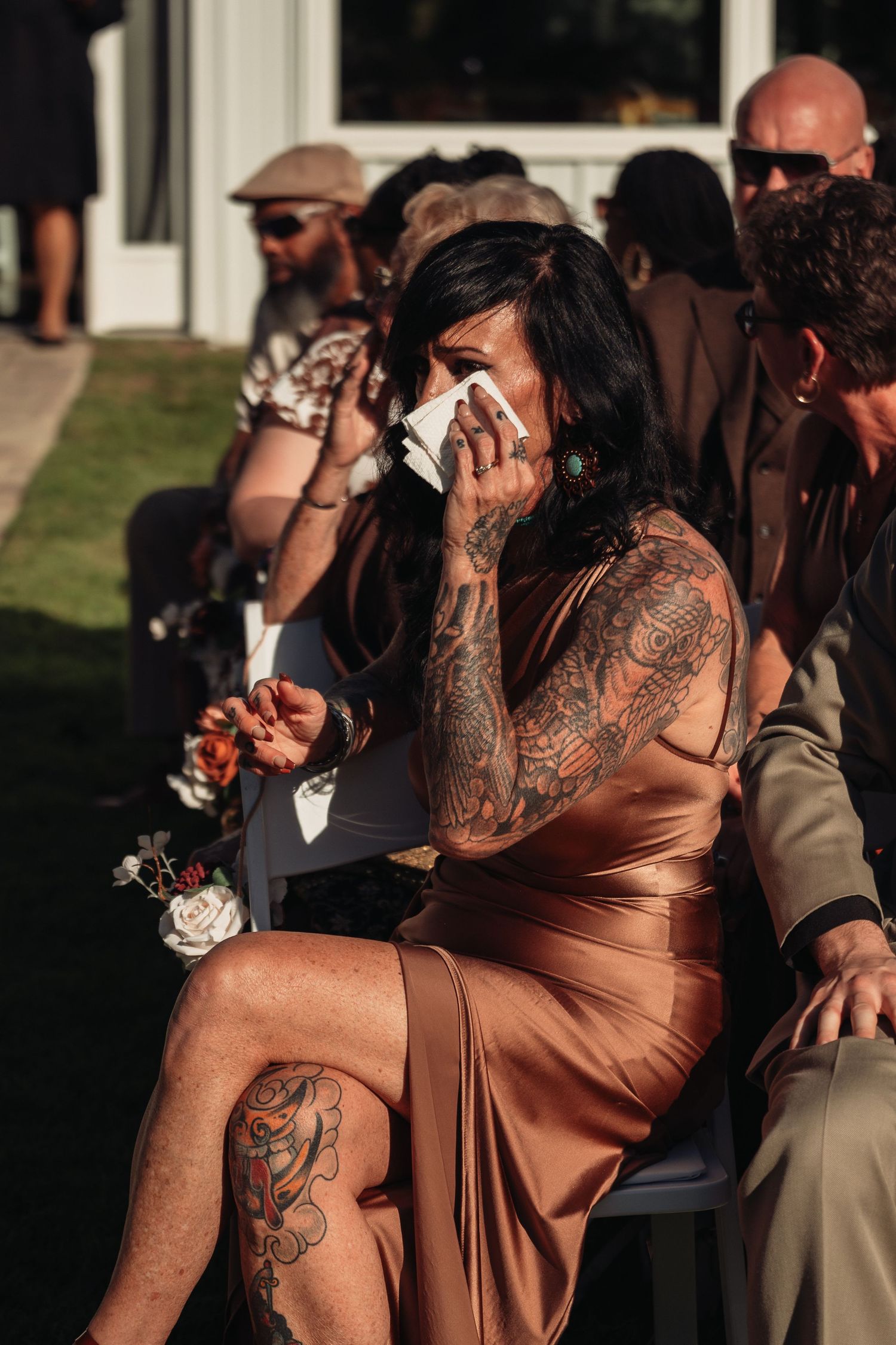 Emotional tattooed wedding guest in copper satin dress wiping tears during outdoor ceremony.