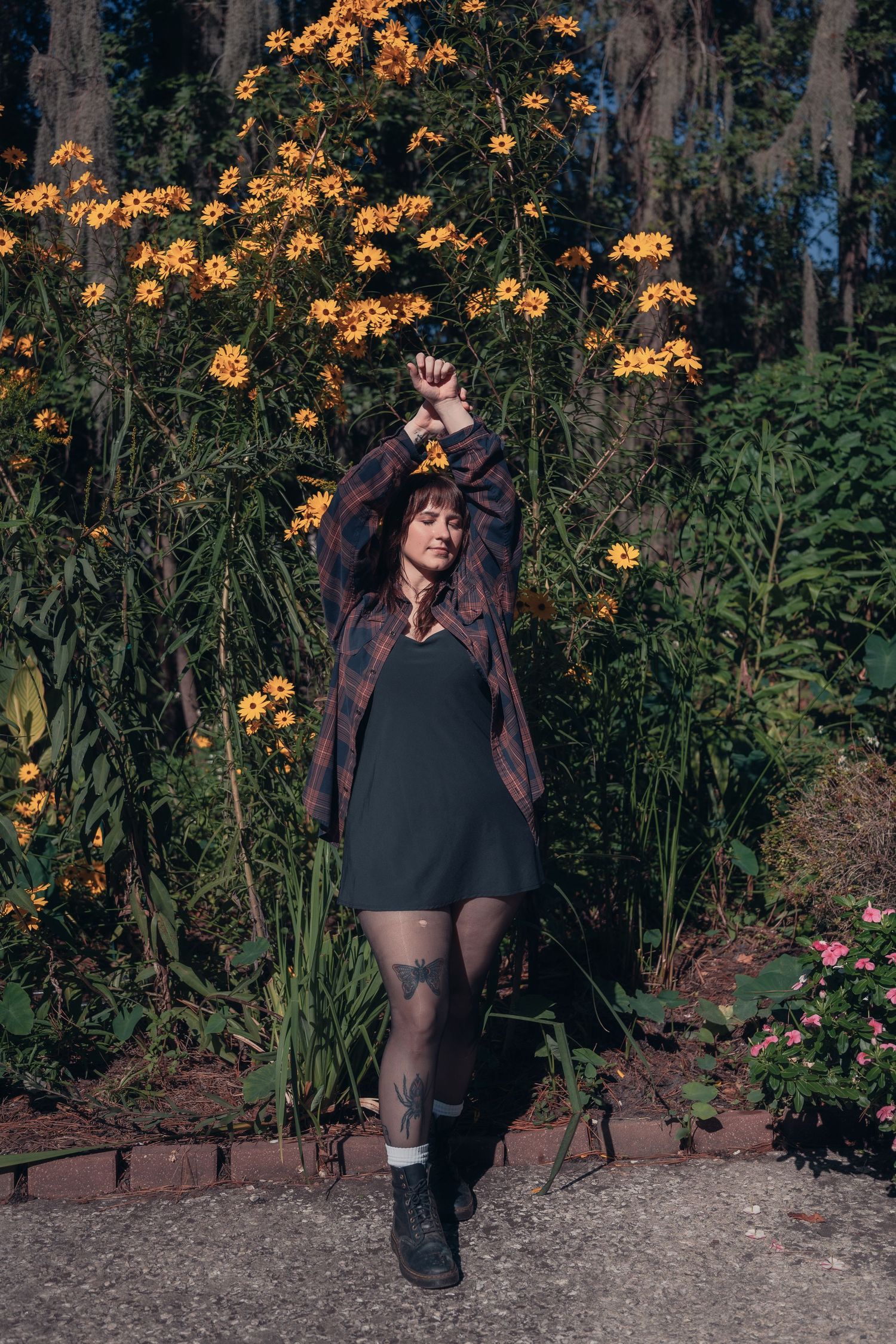 Woman in plaid shirt and black dress poses with raised arms in front of vibrant yellow flowers in a garden.
