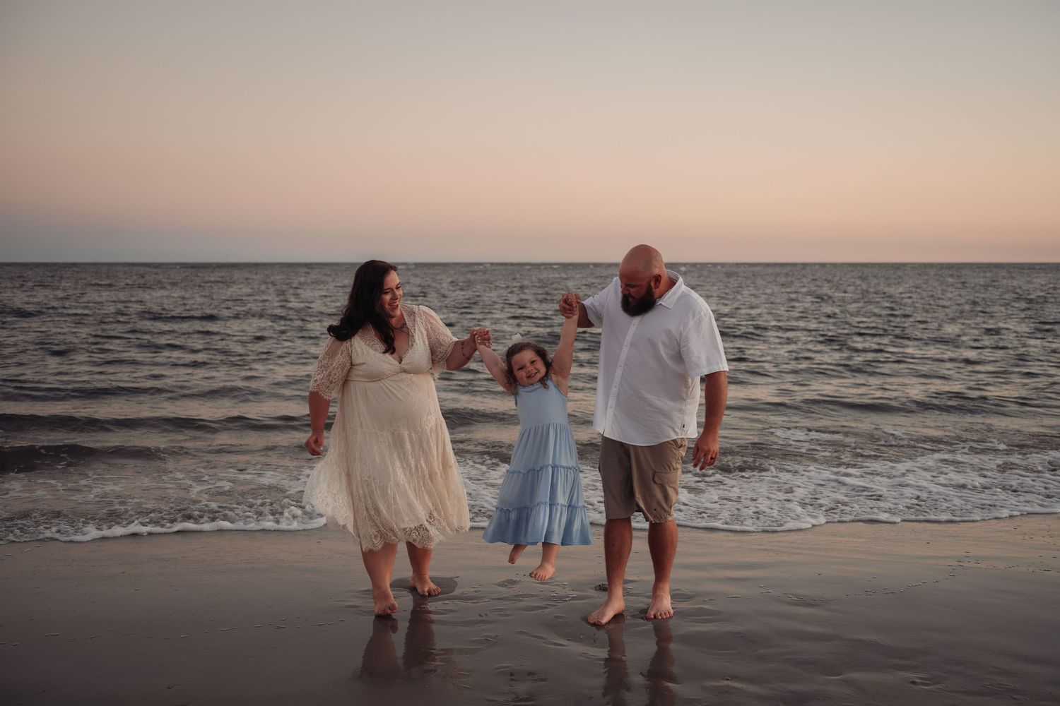 Family of three holding hands and walking along the shoreline of a calm beach at sunset with a young girl swinging between her parents.