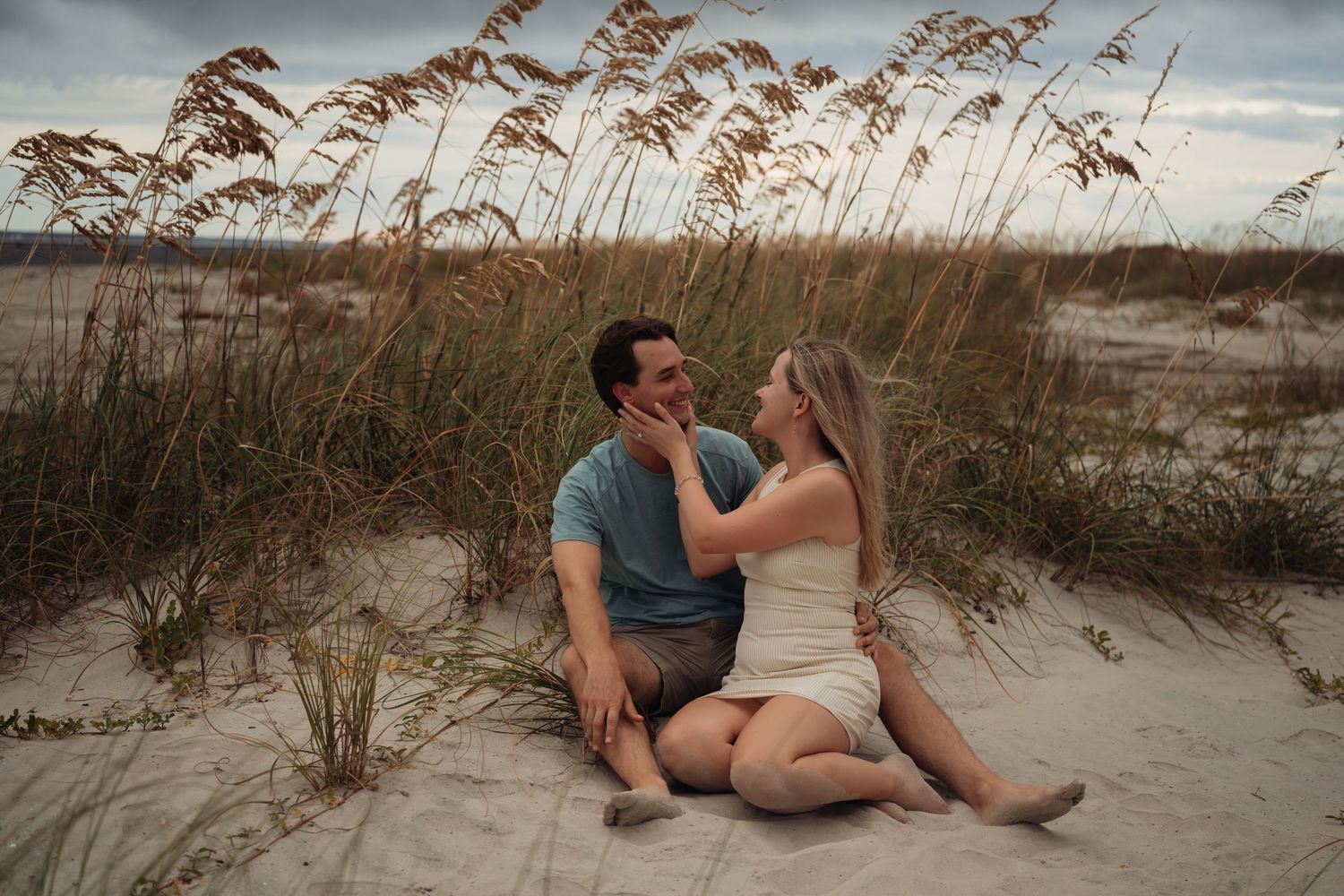 Couple sitting on sandy beach dunes with sea oats, sharing a romantic moment during a moody cloudy day.
