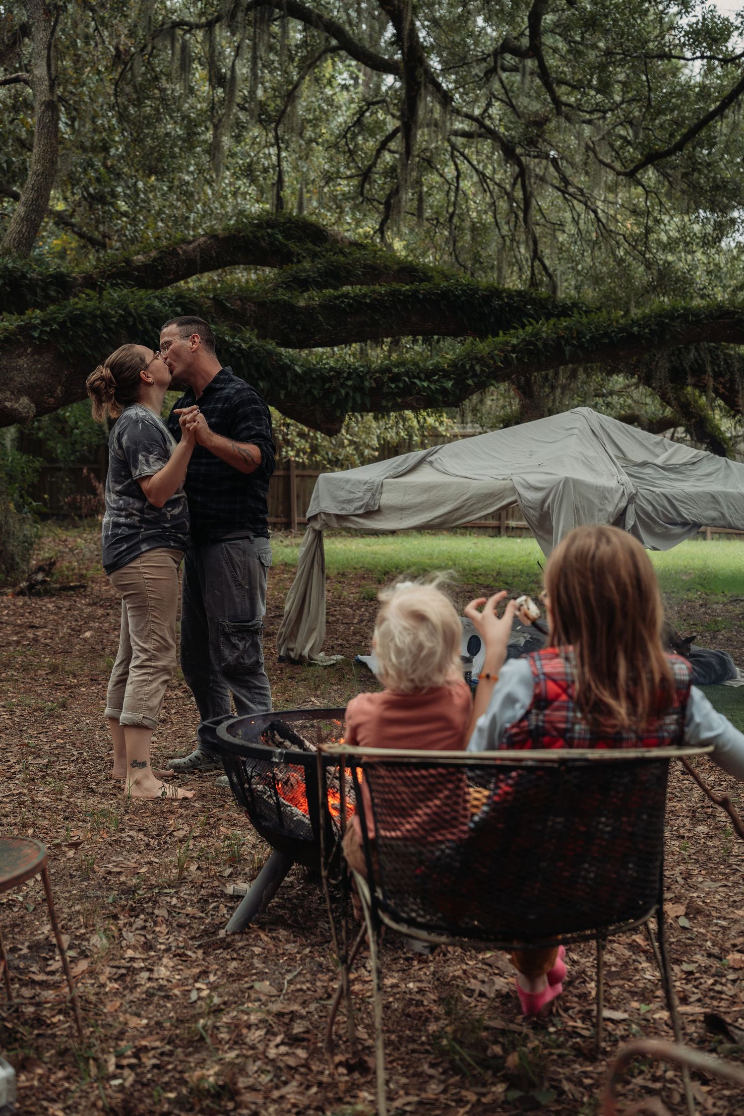 Couple kissing by a campfire in a wooded area while two children sit nearby roasting marshmallows outdoors.