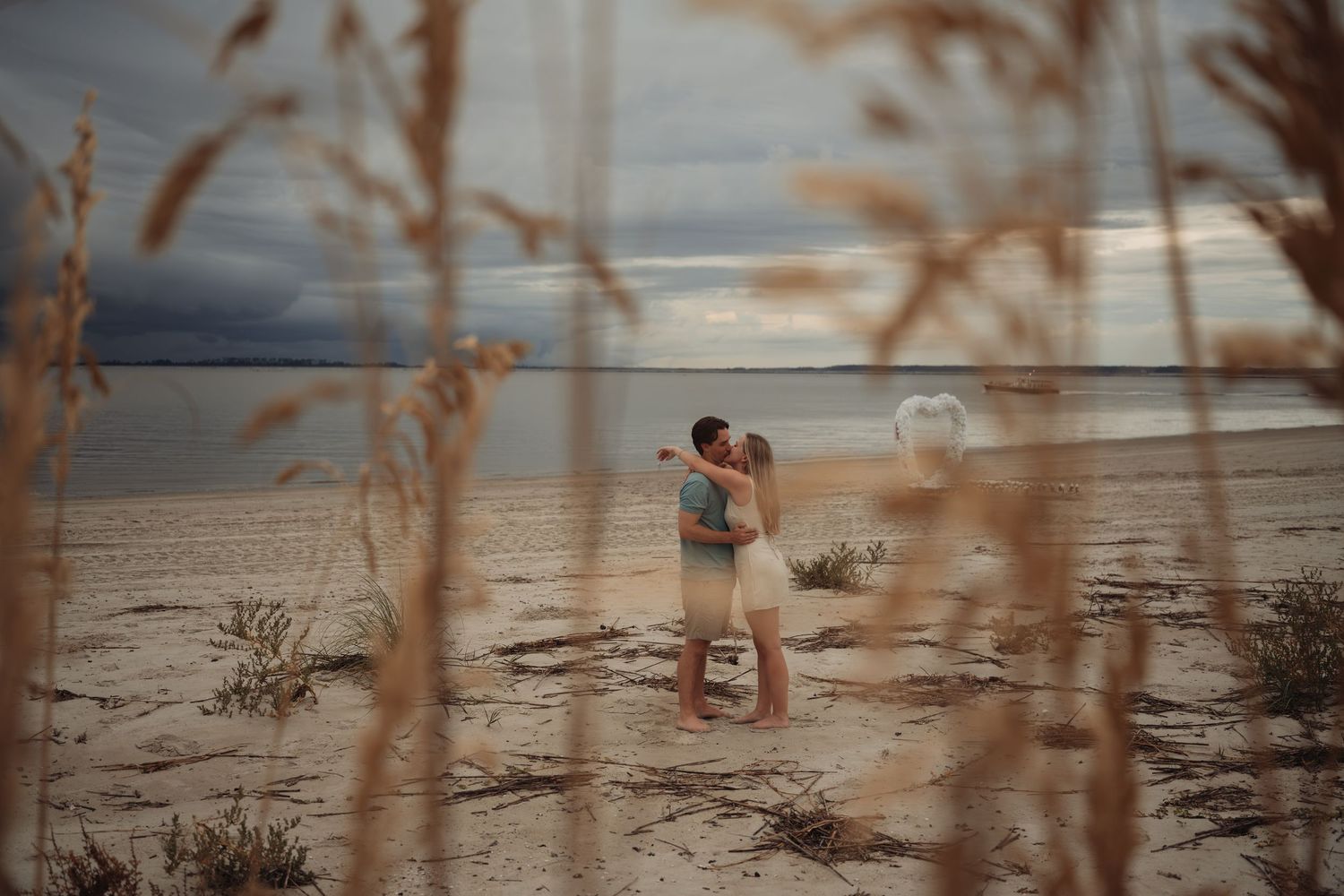 Couple embracing on a sandy beach at dusk, framed by golden sea grass with stormy skies and calm water behind them.