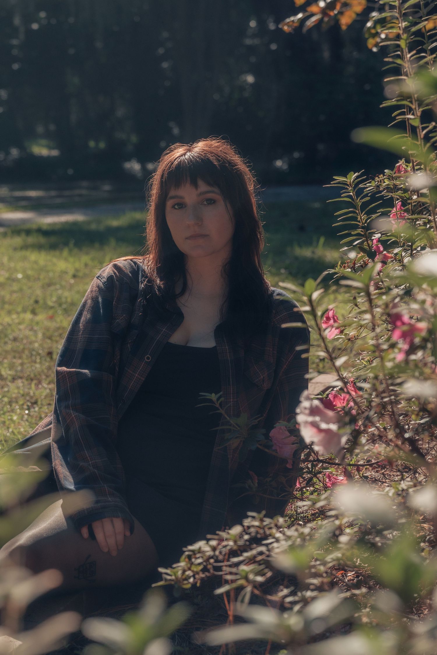 Young woman with dark hair sitting outdoors near pink flowering shrubs in a sunlit park wearing a plaid shirt.