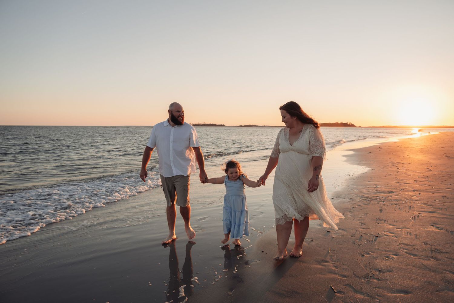 Family of three holding hands and walking along the shoreline of a sandy beach at golden hour sunset with gentle waves.