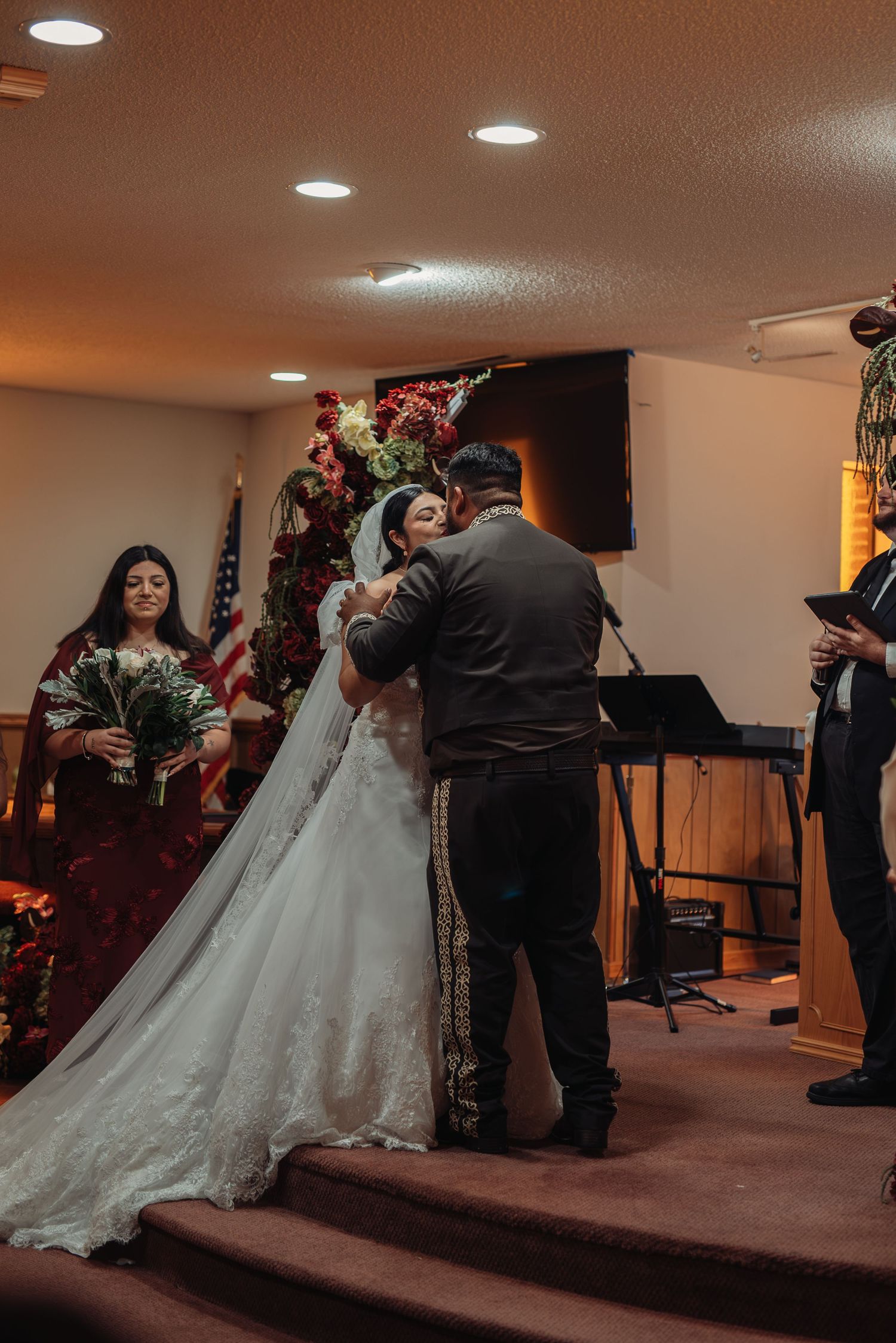 Bride and groom share an emotional embrace at church altar during wedding ceremony with bridesmaid and officiant nearby.
