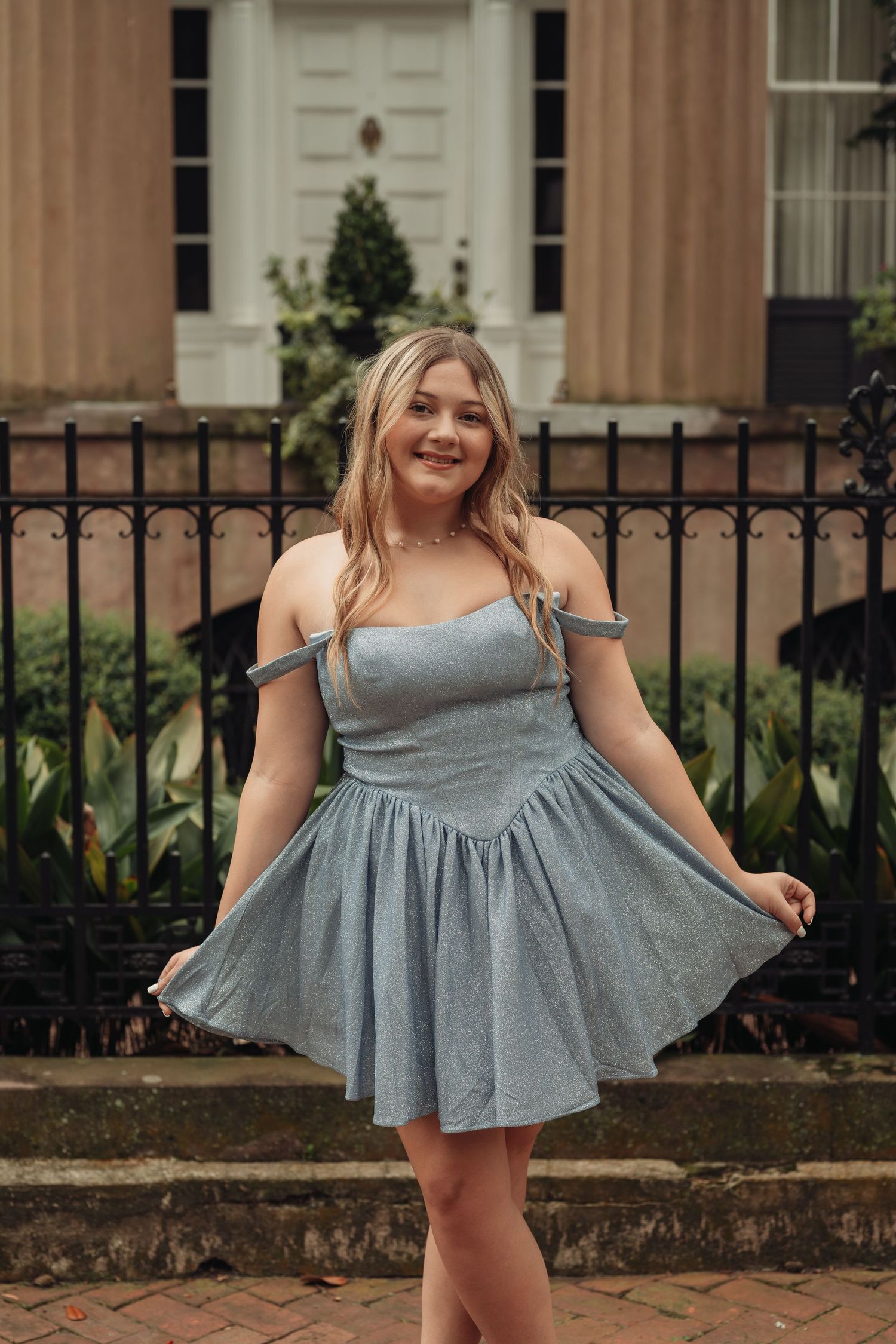 Young woman in light blue off-shoulder fit-and-flare mini dress posing outdoors near iron fence and historic building.