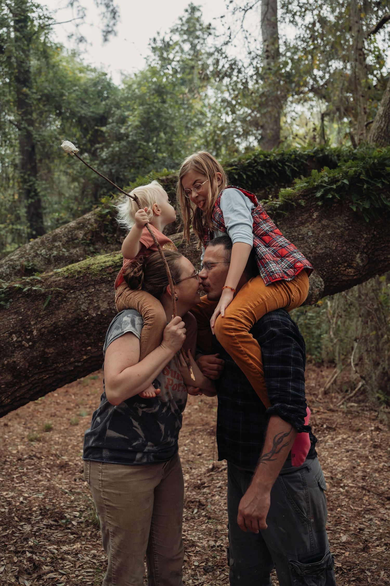 Family of four sharing a kiss in a mossy forest, with two children perched on parents shoulders near a fallen tree.