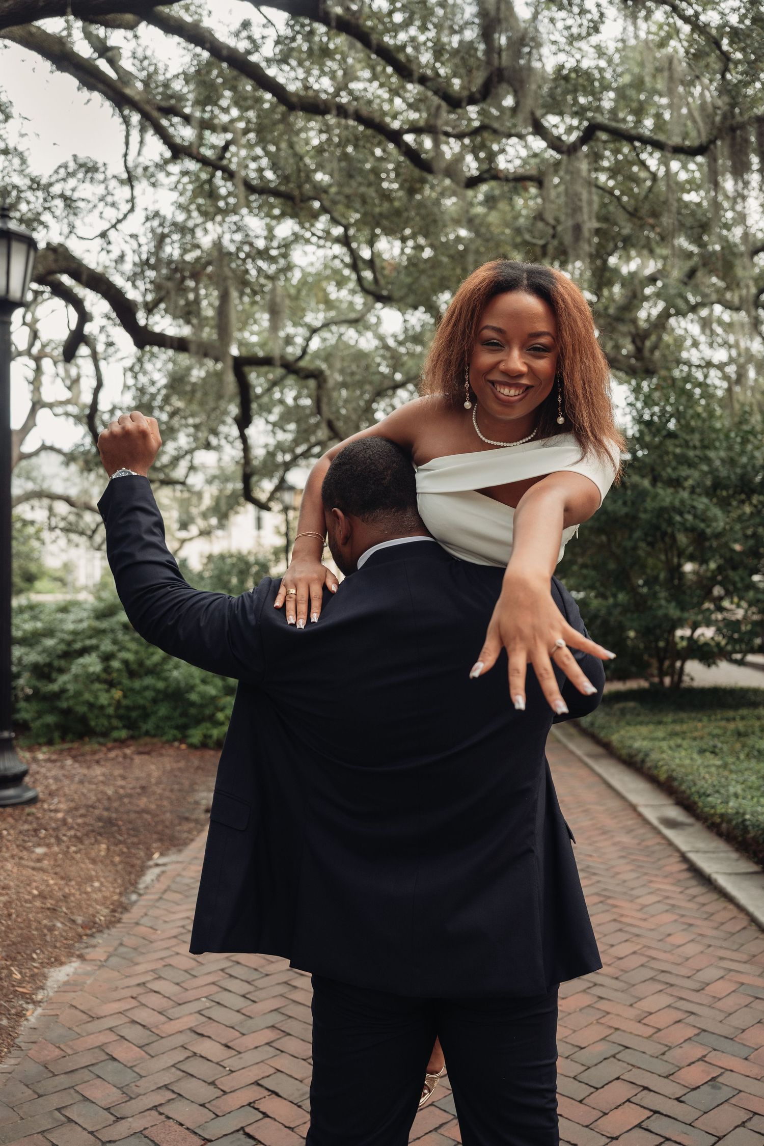 Man lifting smiling woman in white off-shoulder dress along brick pathway under sprawling oak trees.