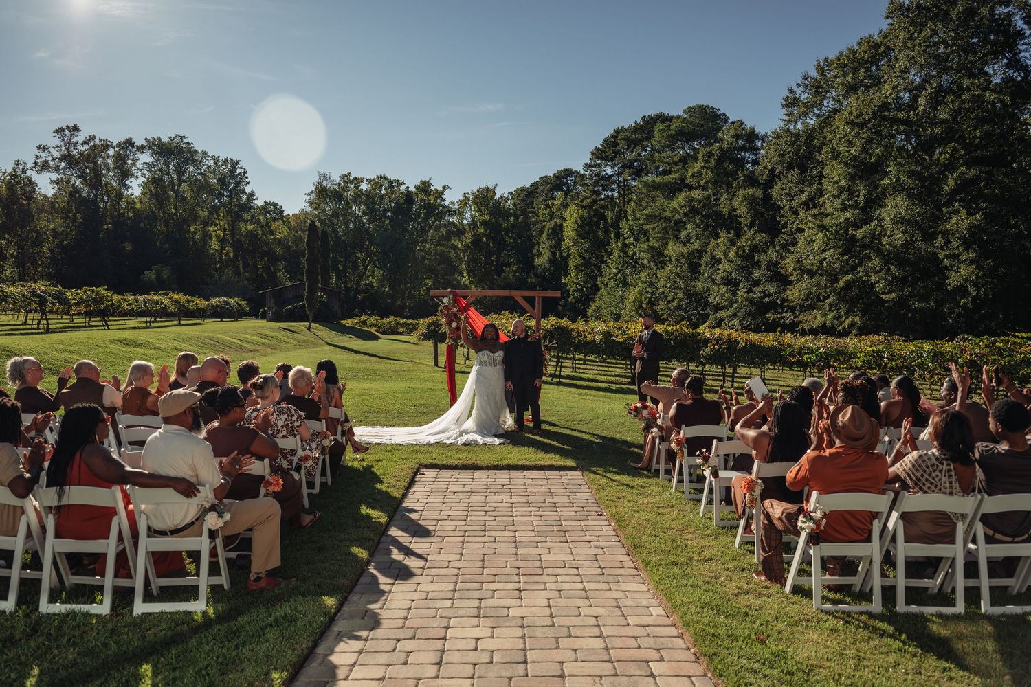 Outdoor wedding ceremony with guests seated along a brick aisle leading to the altar in a vineyard.