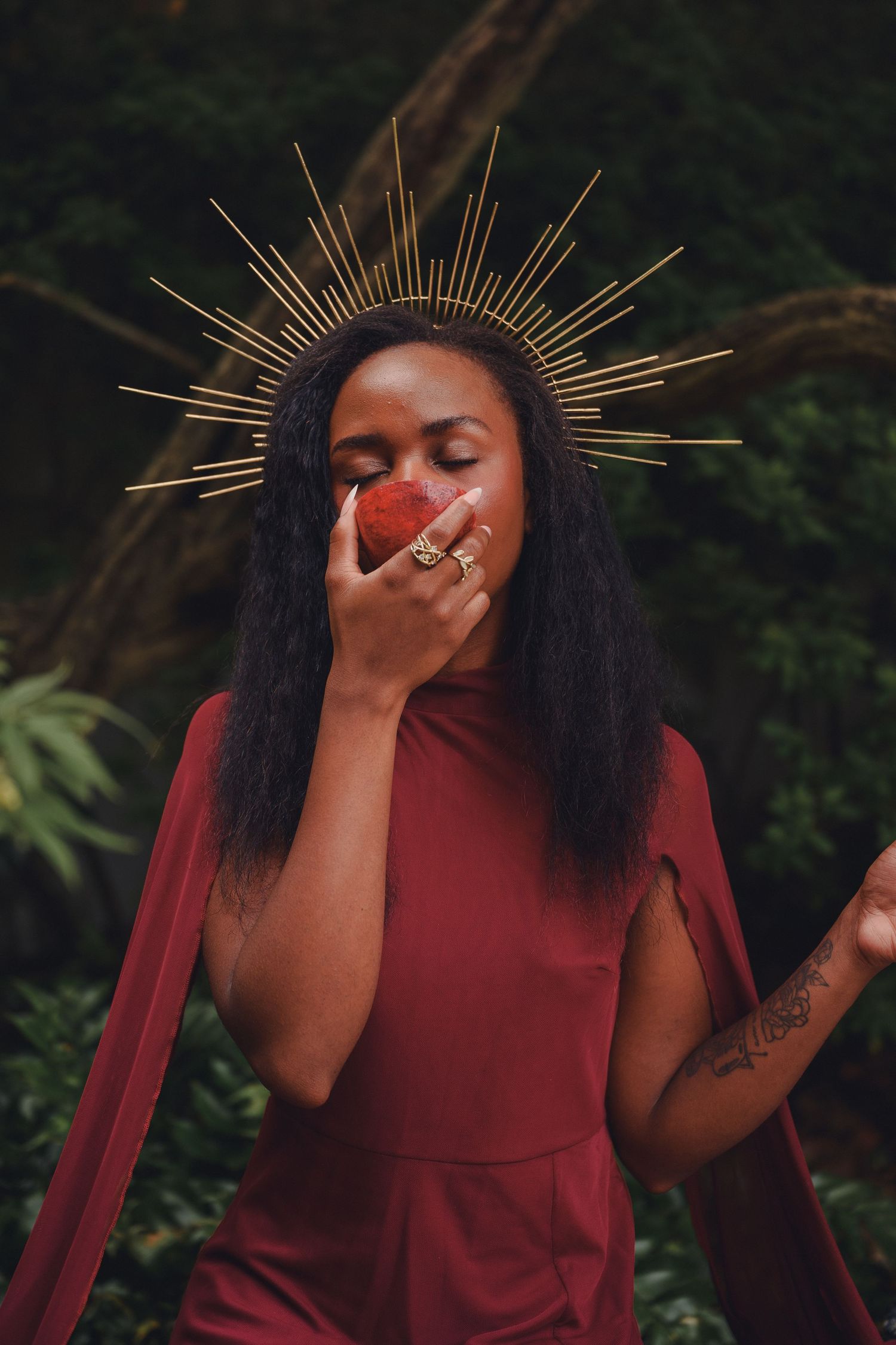 A woman with a gold spiked halo crown in a red dress holds a red flower to her face with eyes closed.
