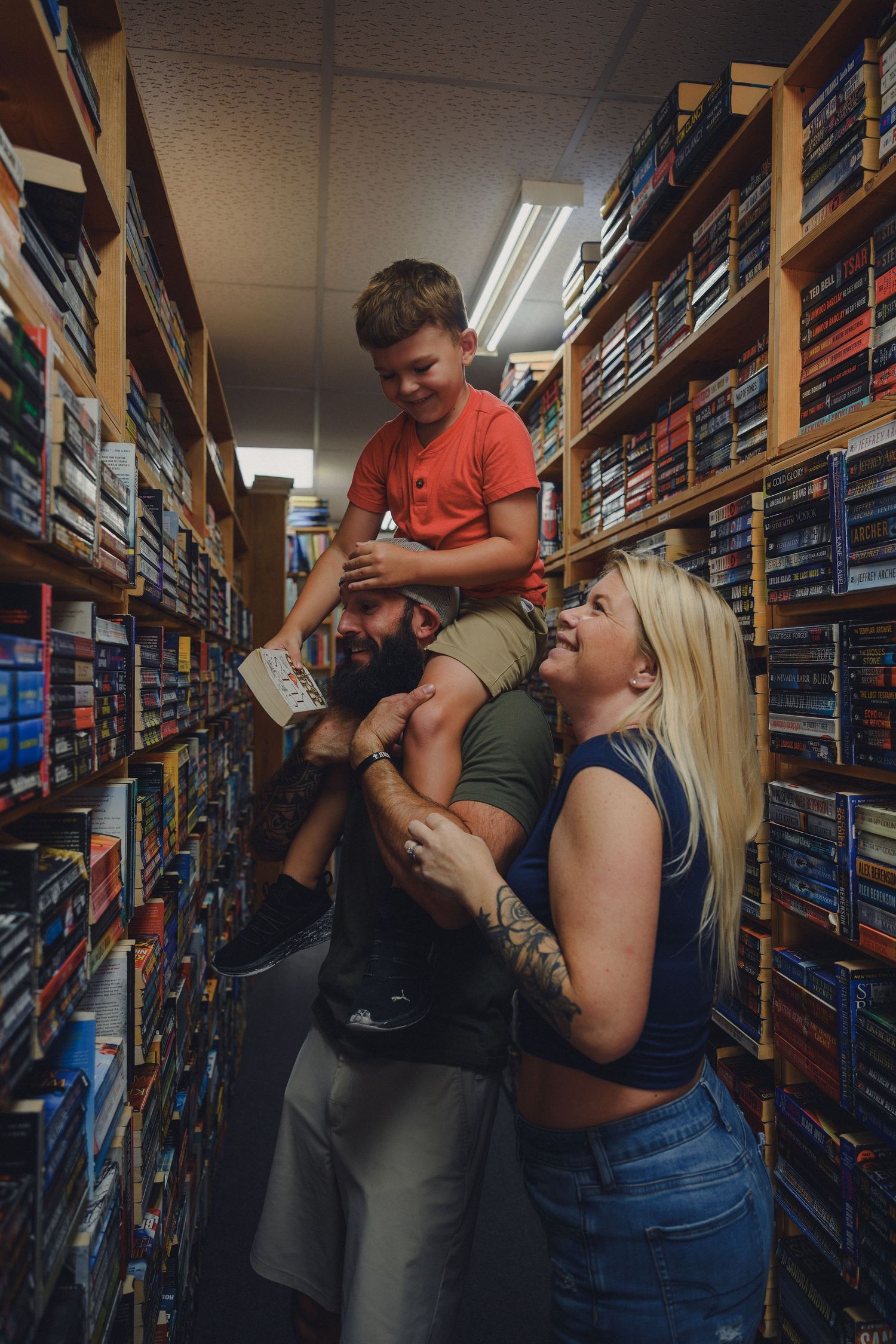 A family smiles in a bookstore aisle, with a young boy sitting on a bearded man's shoulders while a blonde woman laughs beside them.