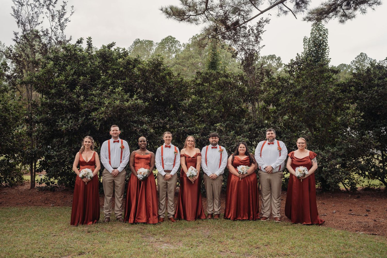 Wedding party of bridesmaids in rust-colored dresses and groomsmen in suspenders standing in a row on a grassy lawn with trees behind.
