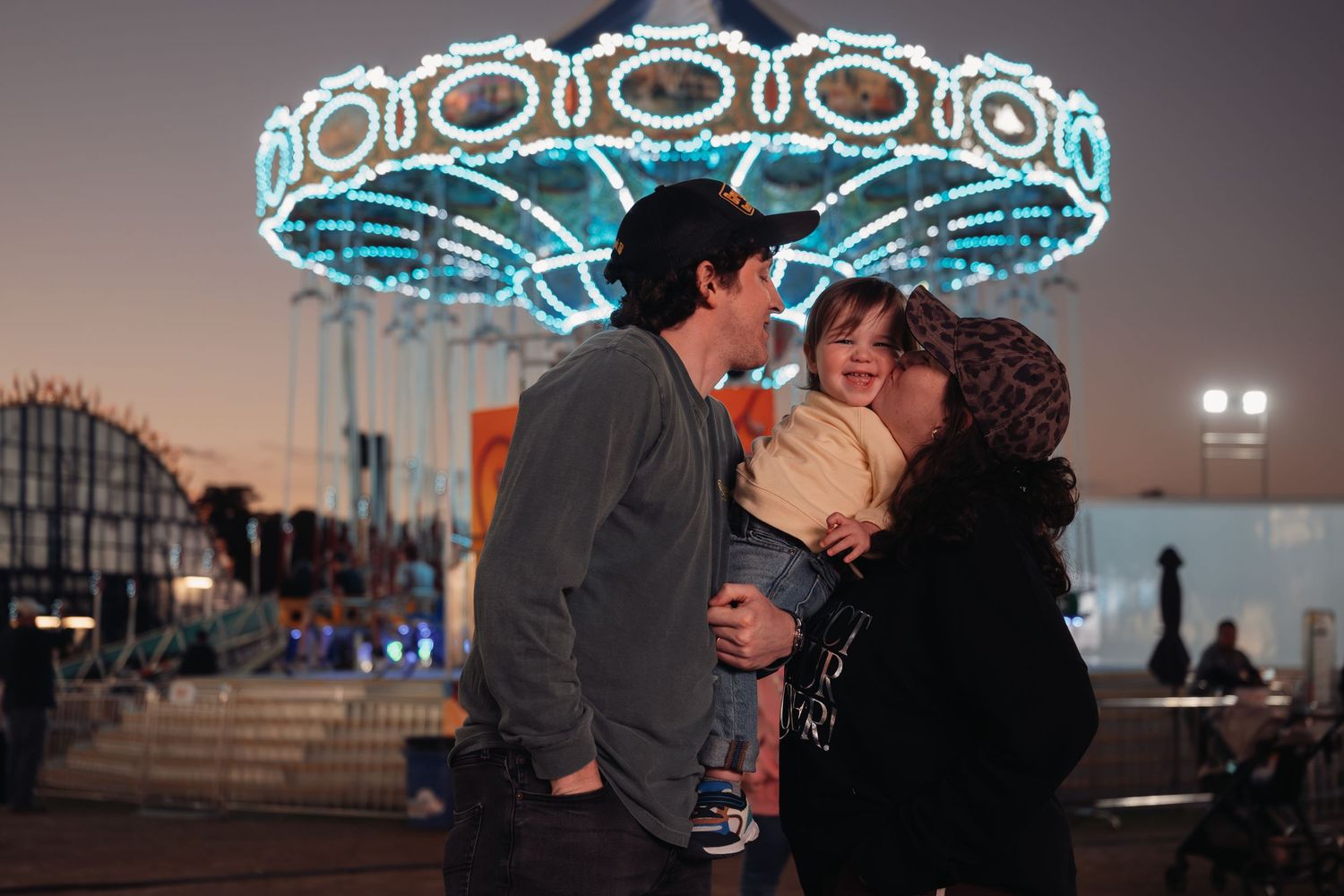 A couple holds a toddler at an amusement park at dusk, with a glowing illuminated carousel and carnival rides in the background.