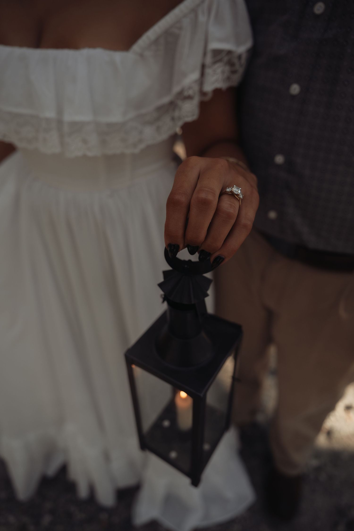 Close-up of a couple holding a black lantern with a lit candle, woman wearing an engagement ring and white off-shoulder dress.