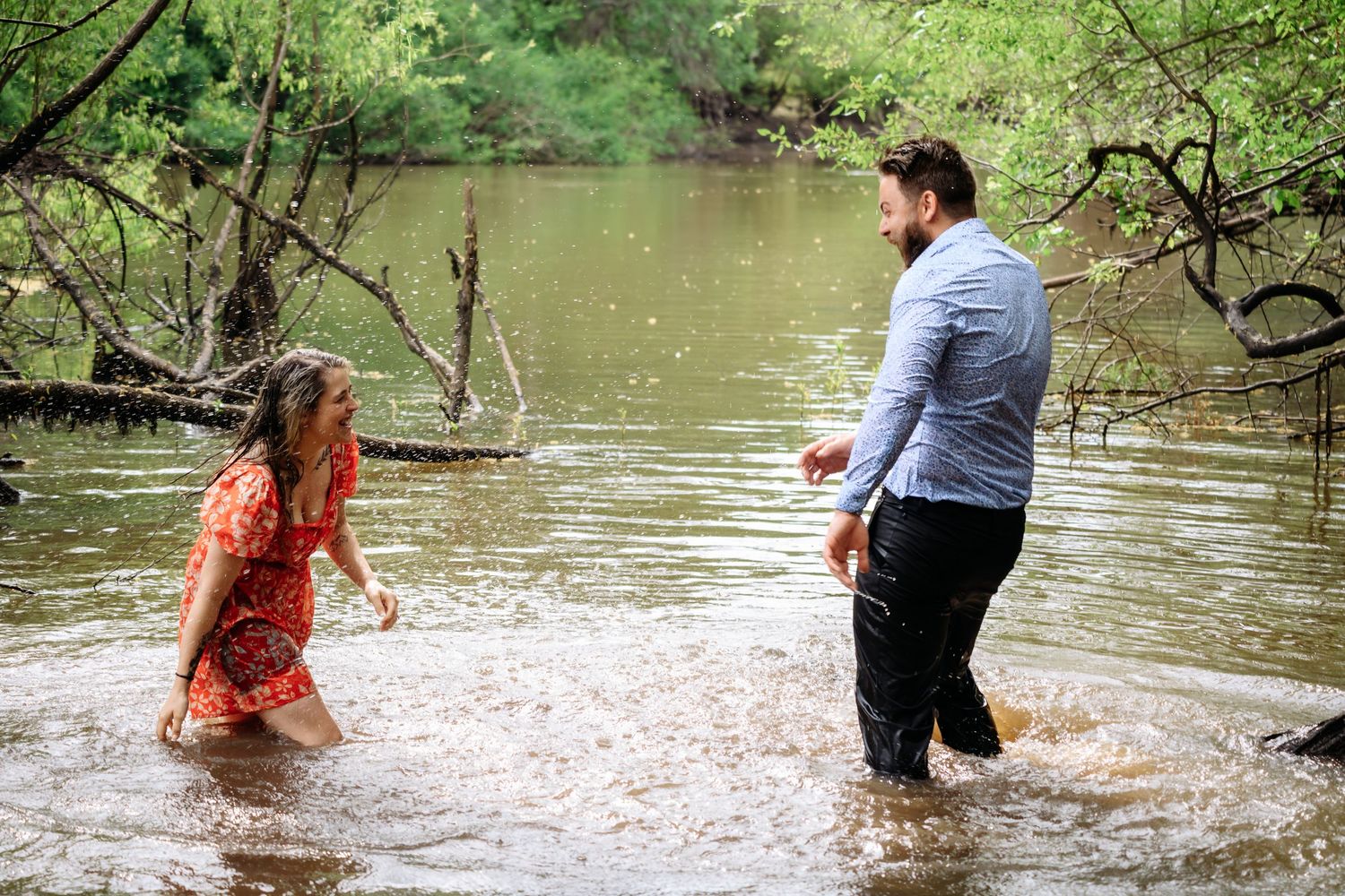 Couple wading through shallow river water surrounded by lush green trees during outdoor photo session.