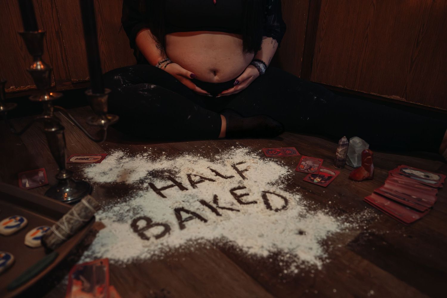 Flour spelling Half Baked on wooden floor with pregnant belly, candles, and tarot cards nearby.