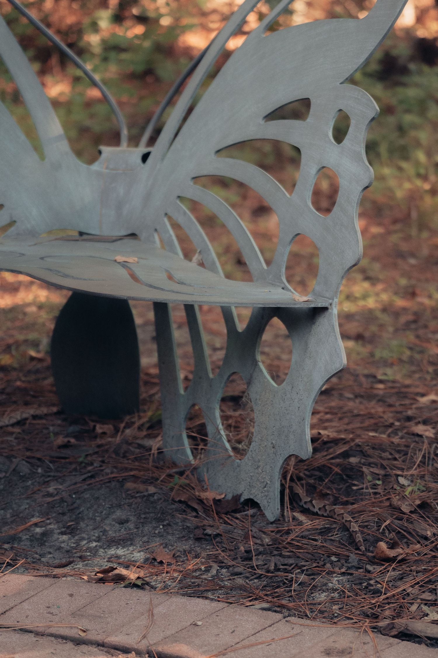 Artistic metal butterfly-shaped bench with intricate cutout design, set outdoors among pine needles and natural surroundings.