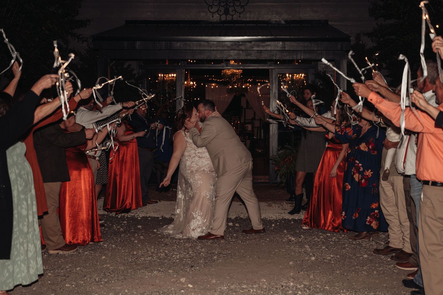 Bride and groom embrace as wedding guests form an arch with sparklers at a nighttime outdoor reception.