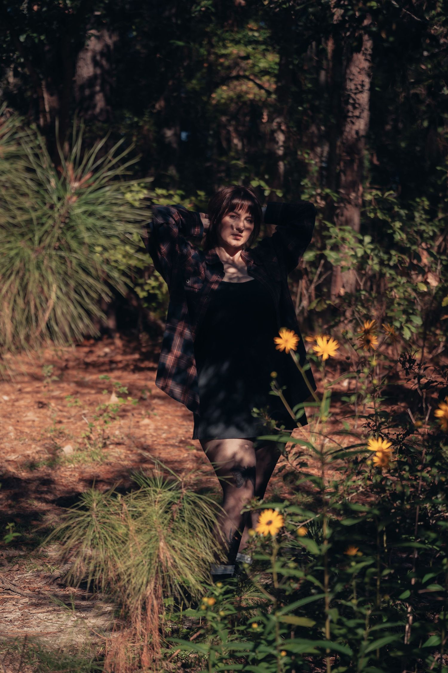Young woman in black dress posing among yellow wildflowers and tall trees in a sunlit forest setting.