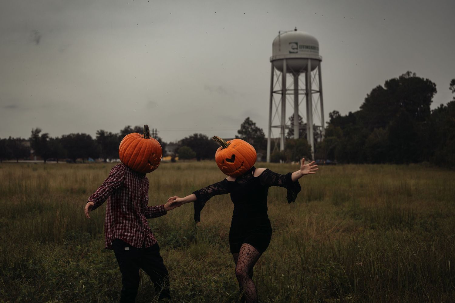 Two people wearing pumpkin heads dance in a field with a water tower in the background on a cloudy day.