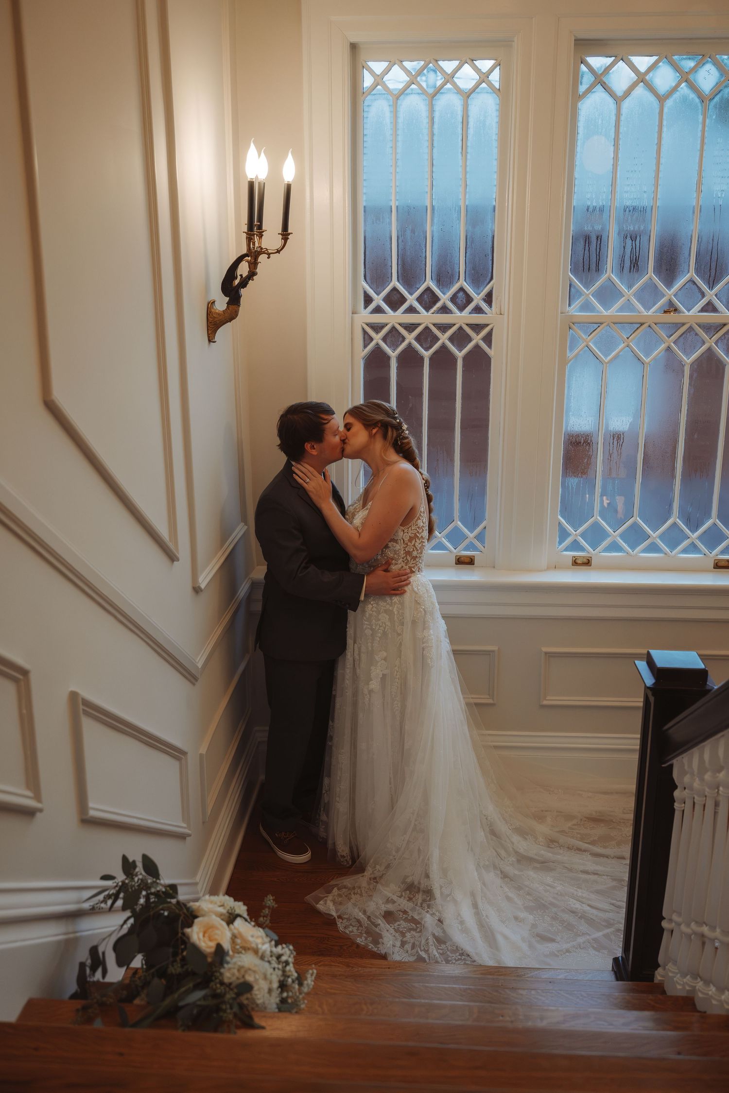 Bride and groom share a romantic kiss on a staircase landing by a latticed window in an elegant venue.