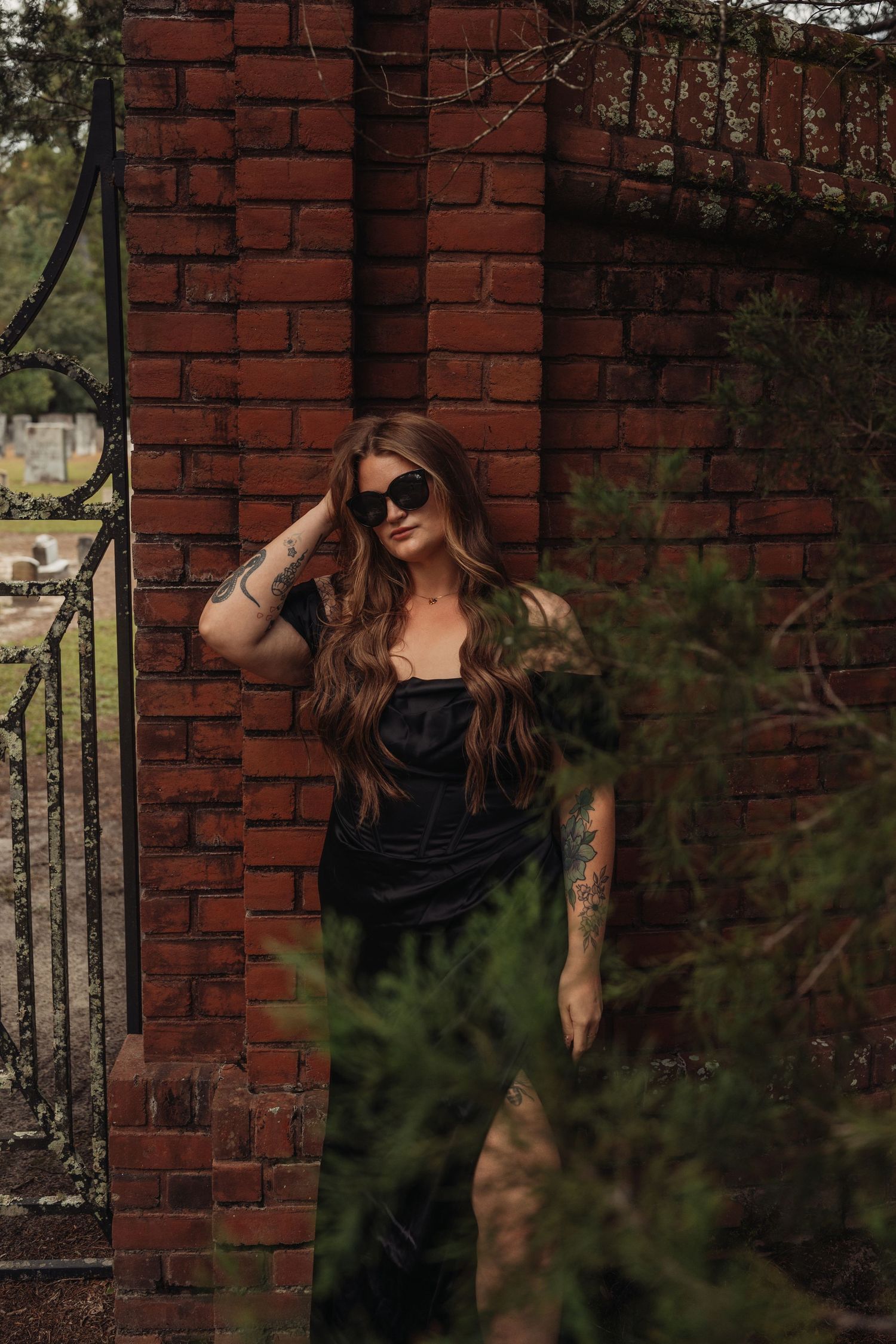 Woman in black dress and sunglasses with tattoos posing against a red brick wall outdoors.