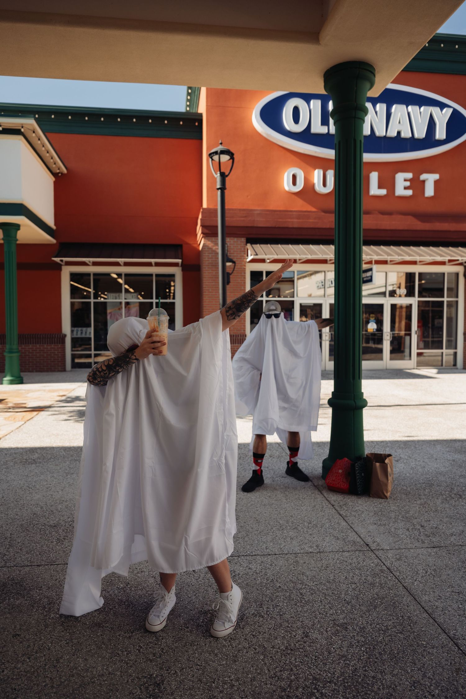 Person in a white ghost costume reaches up a green lamppost outside an Old Navy Outlet store.