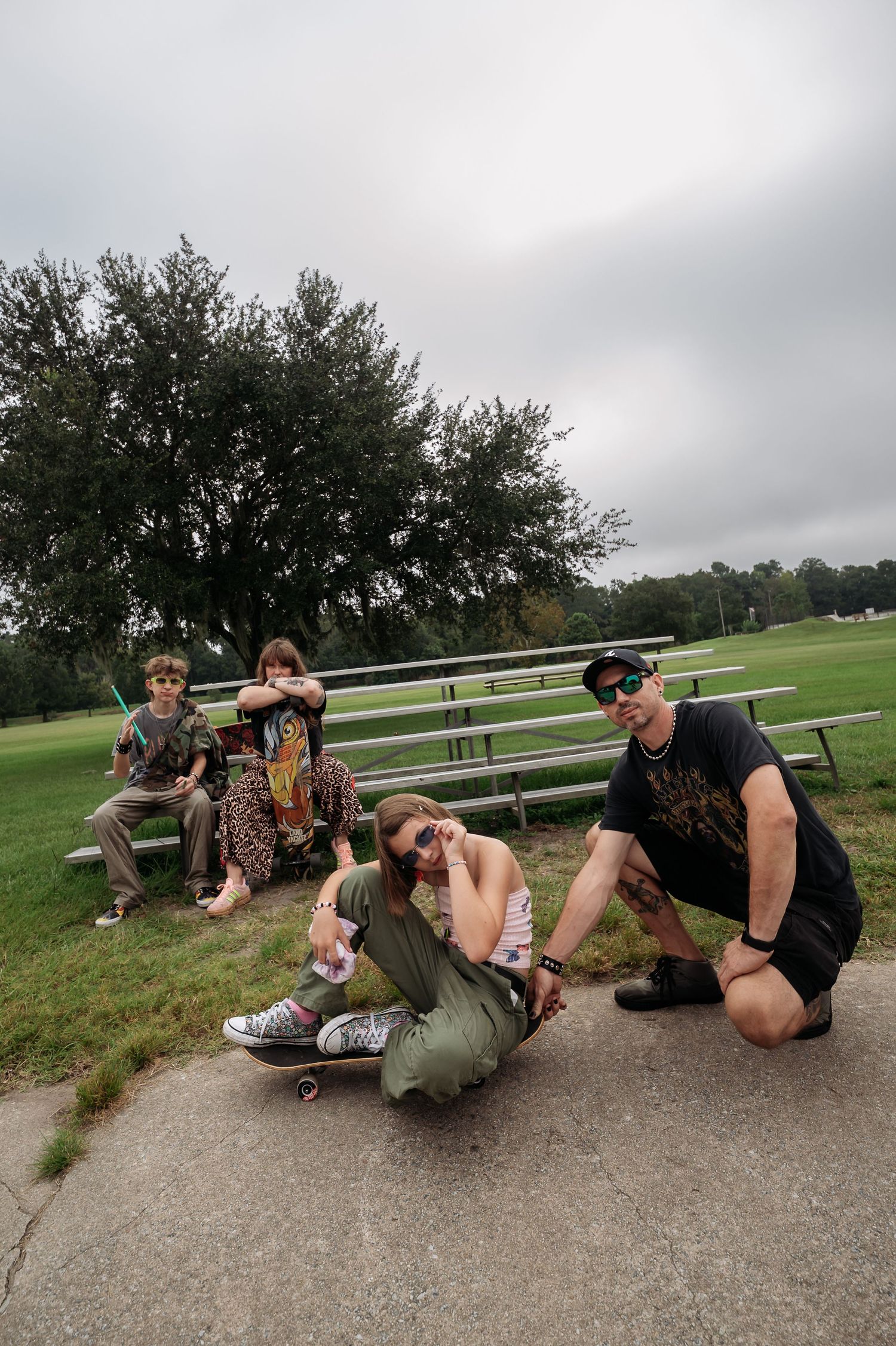 Four people pose casually at a grassy park with bleachers, one woman sitting on a skateboard and a man in sunglasses crouching beside her.