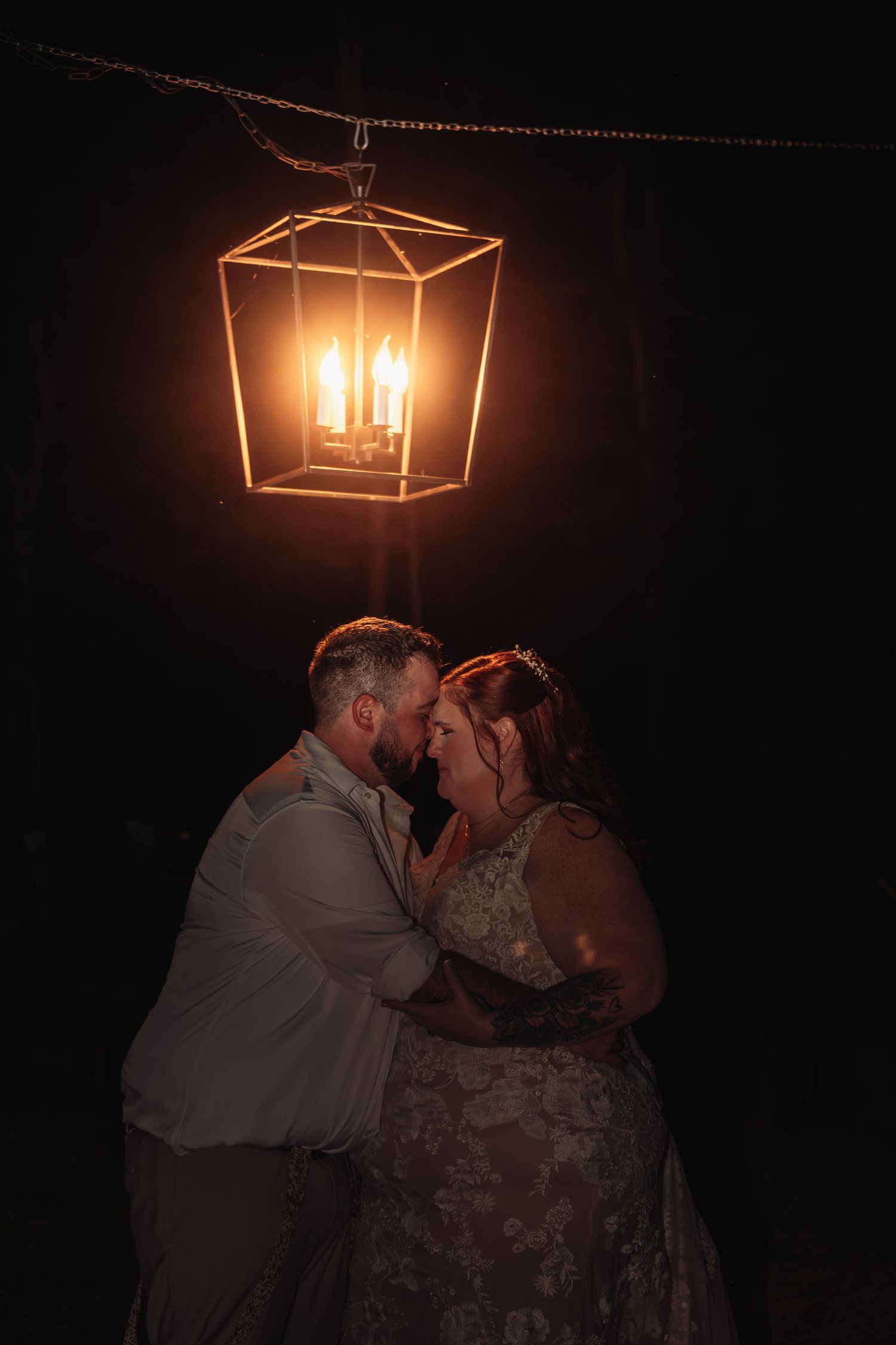Bride and groom share an intimate embrace at night, foreheads touching, beneath a glowing candlelit lantern hanging in the dark.