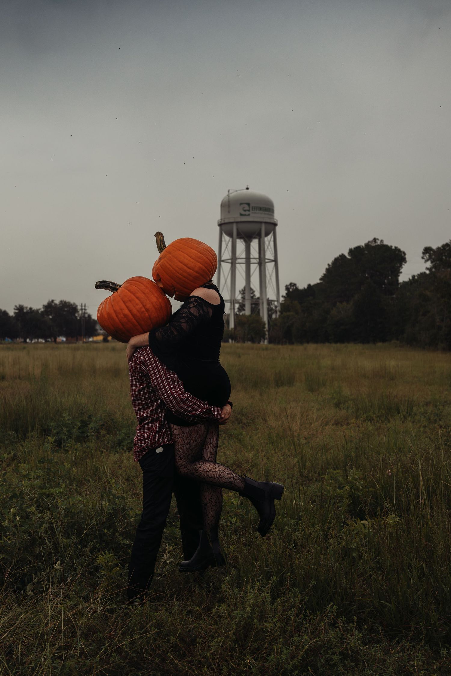 Couple kissing in a field holding pumpkins over their heads with a water tower in the background on a cloudy day.