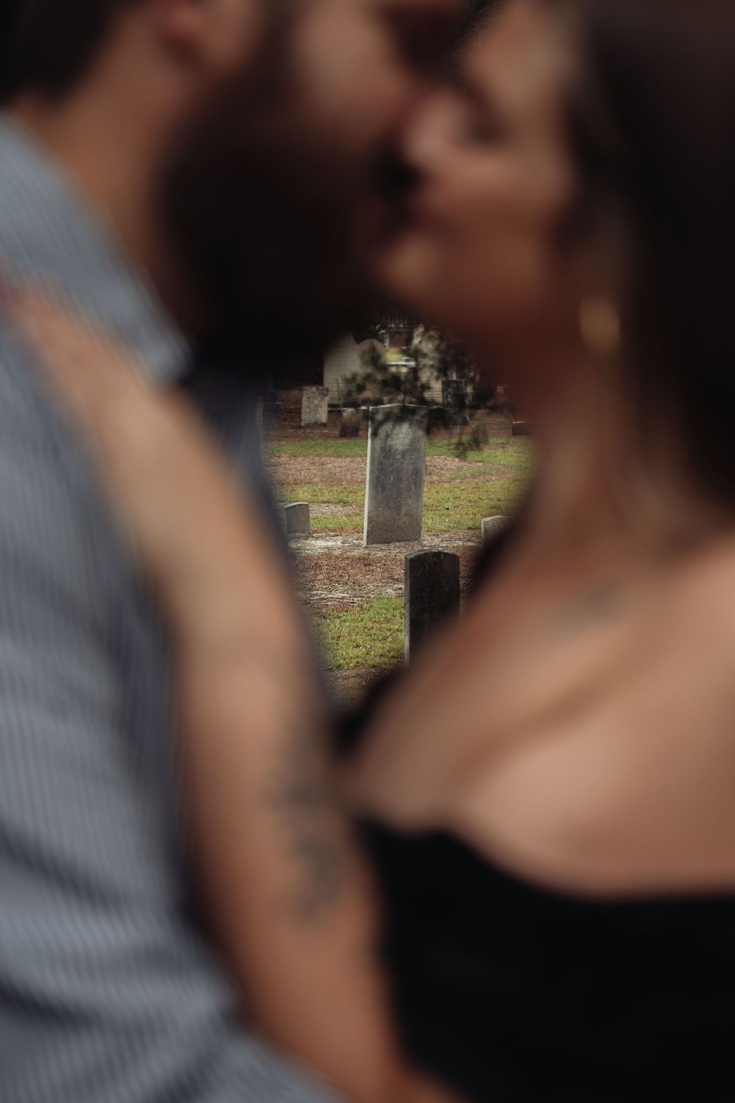Couple sharing a romantic kiss in a cemetery, with gravestones softly blurred in the background.