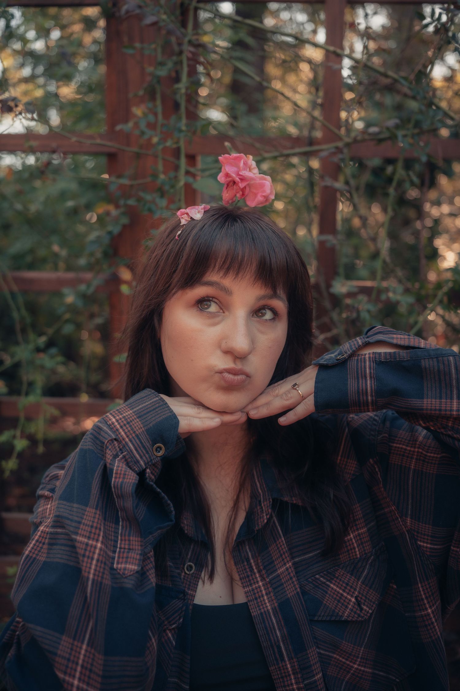 Young woman with pink flower in hair, wearing plaid shirt, making a kissing face outdoors near rustic trellis.