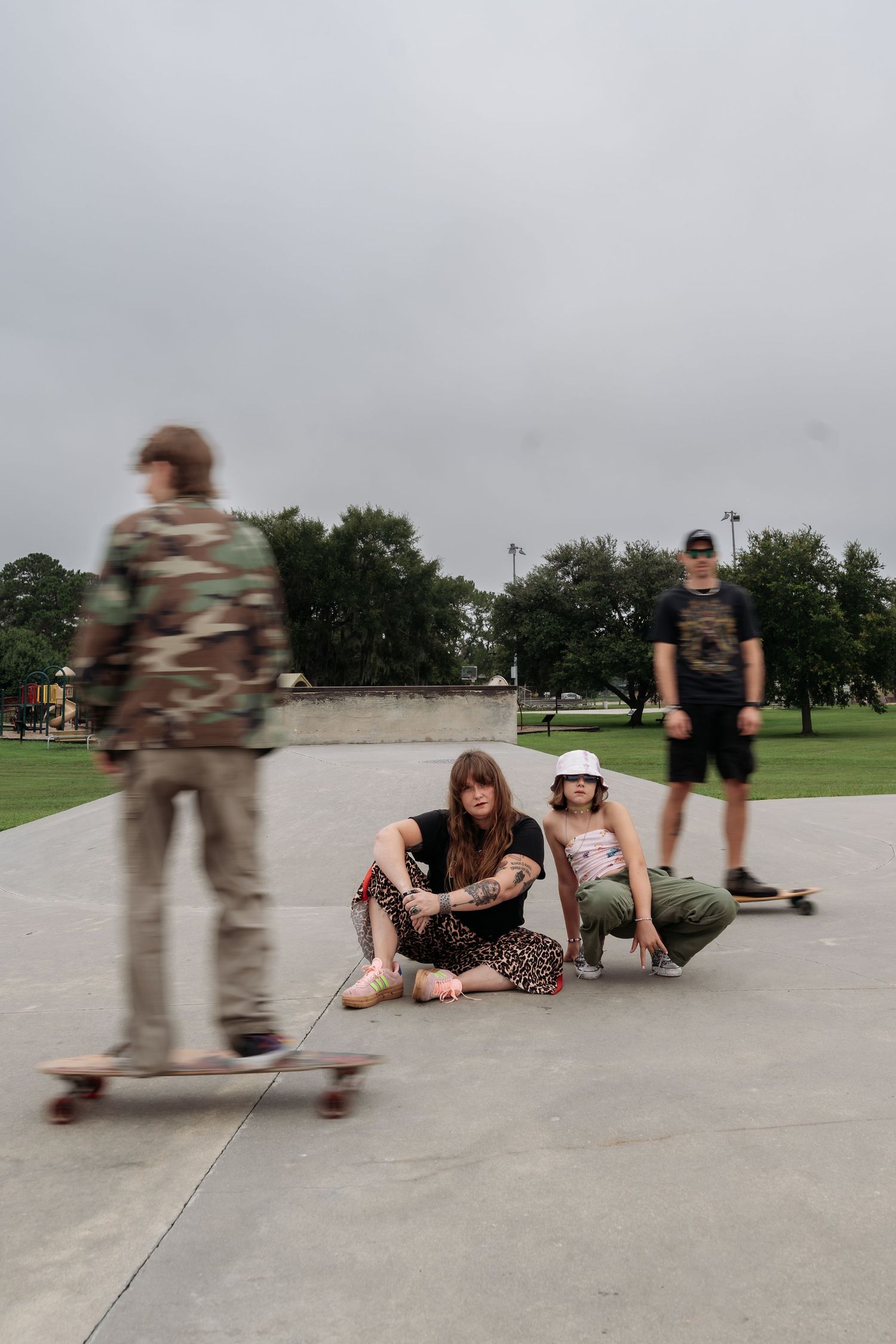 Three friends at a concrete skate park on an overcast day, two sitting casually while one skateboards in a blurred camo jacket.