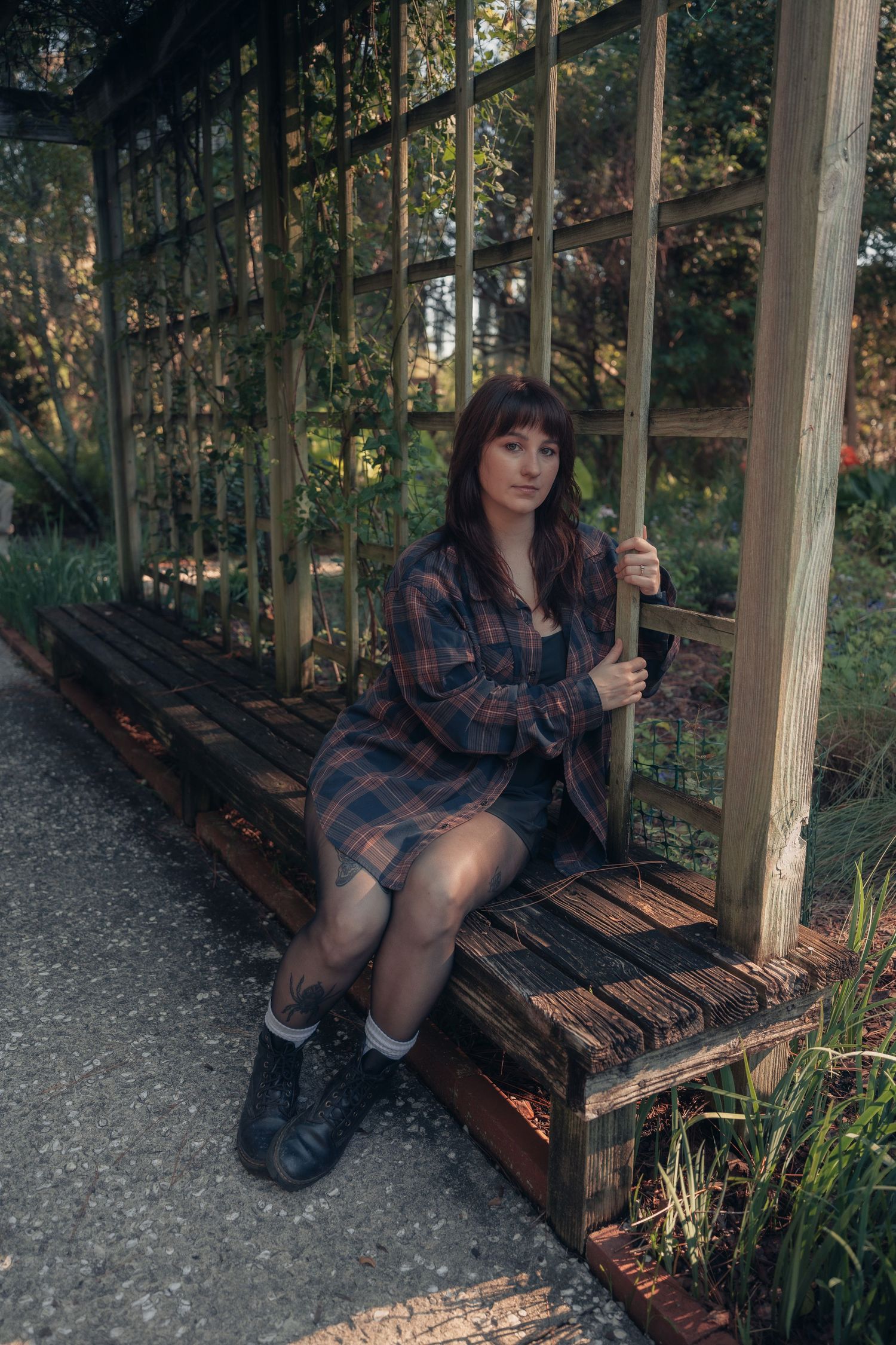 Young woman in plaid shirt dress and black boots sitting on wooden bench in a lush garden pergola.