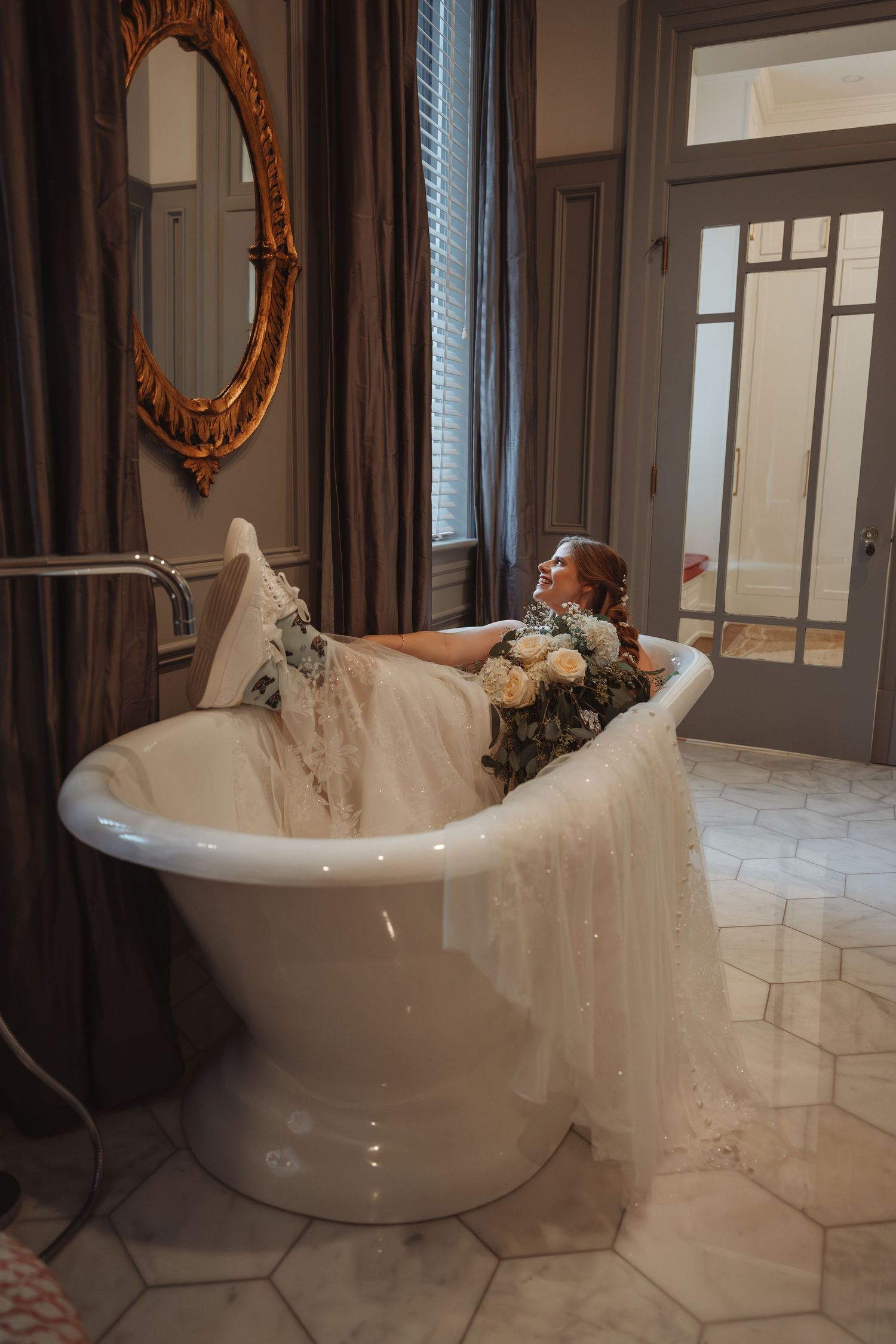 Bride in wedding gown sitting in a freestanding clawfoot bathtub holding a floral bouquet in an elegant bathroom.