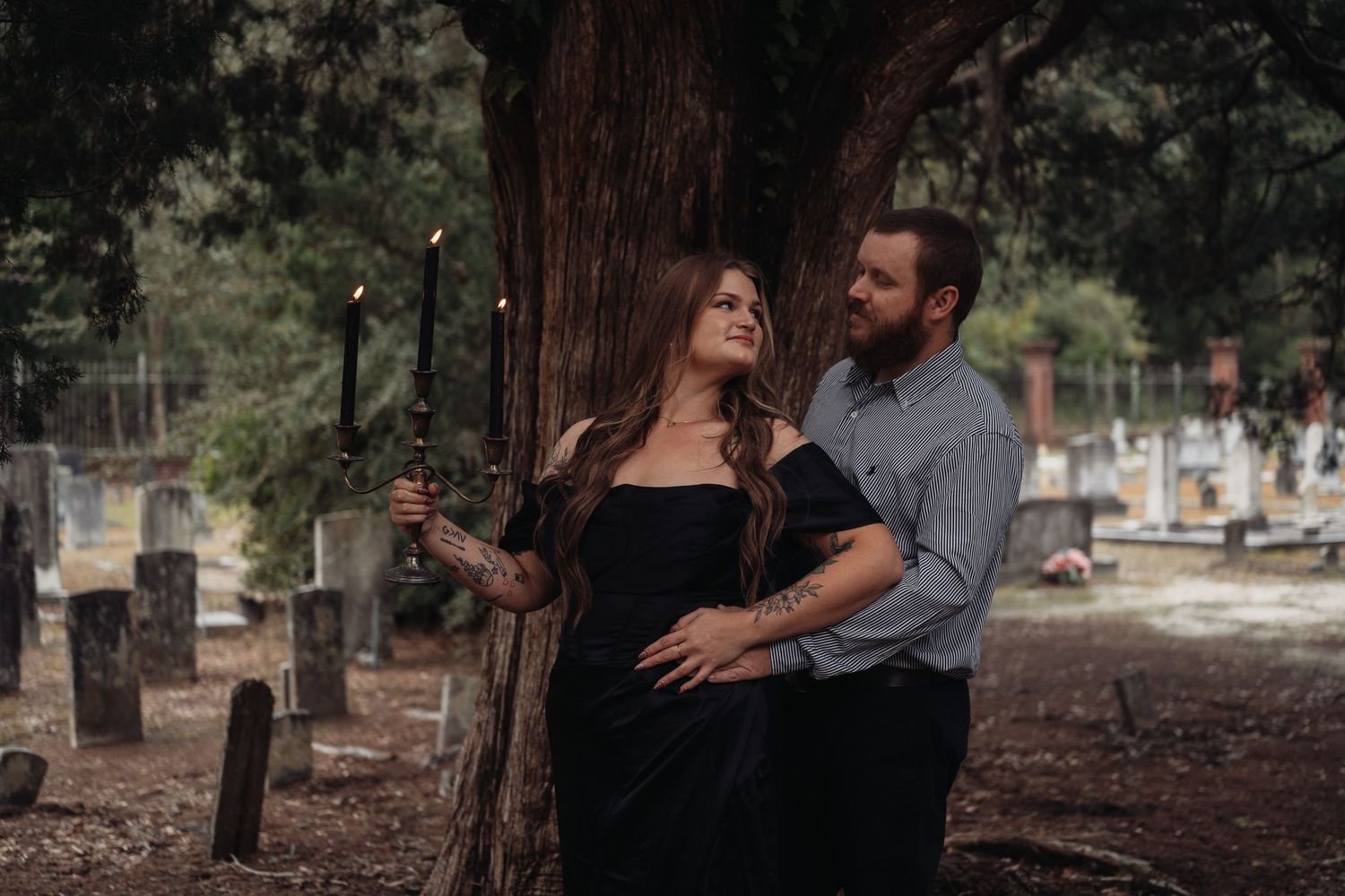 Couple embracing romantically by a tree in a historic cemetery, woman in black dress holding candles.