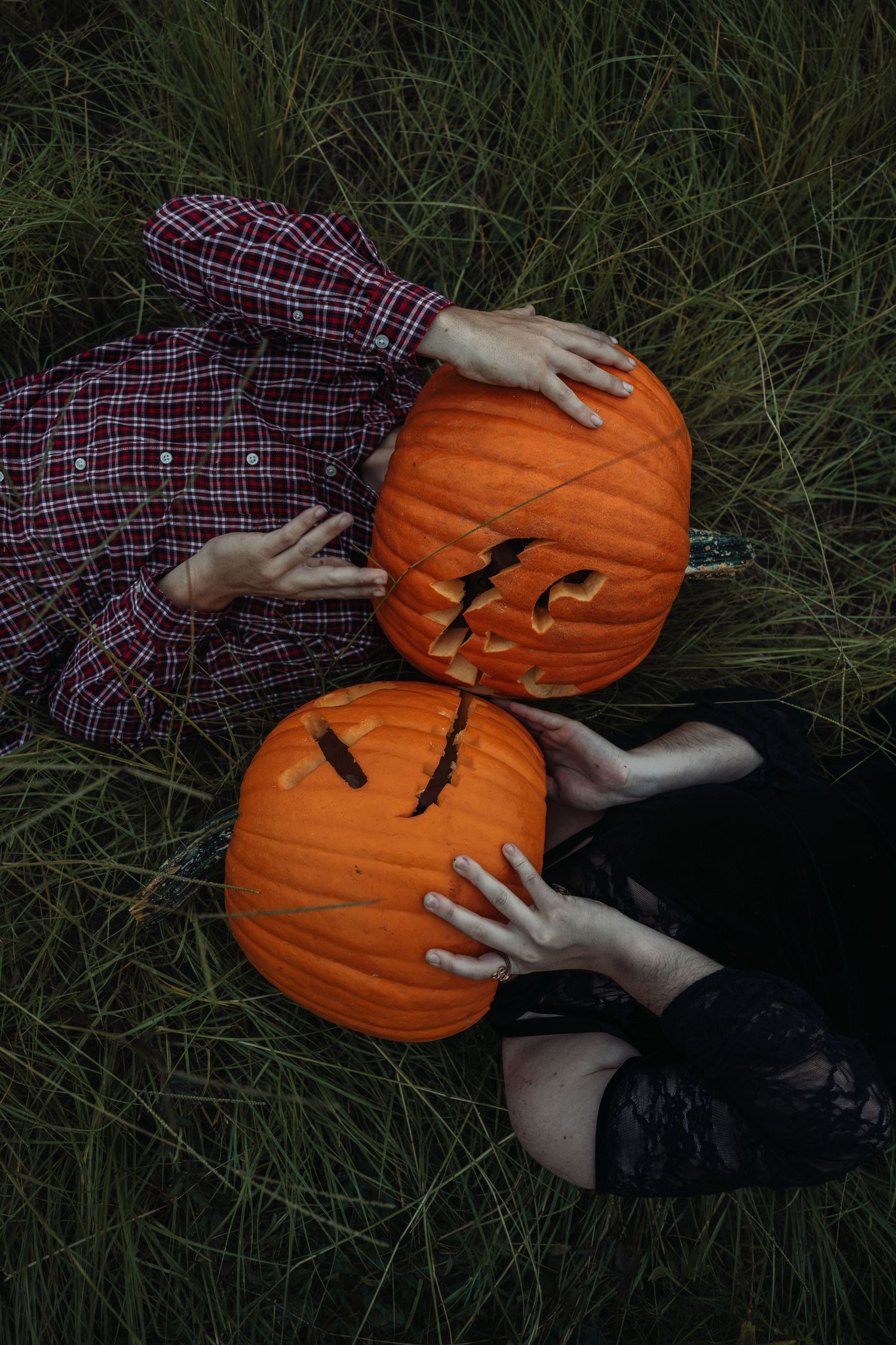 Two people lying in grass holding carved Halloween jack-o-lantern pumpkins over their faces.