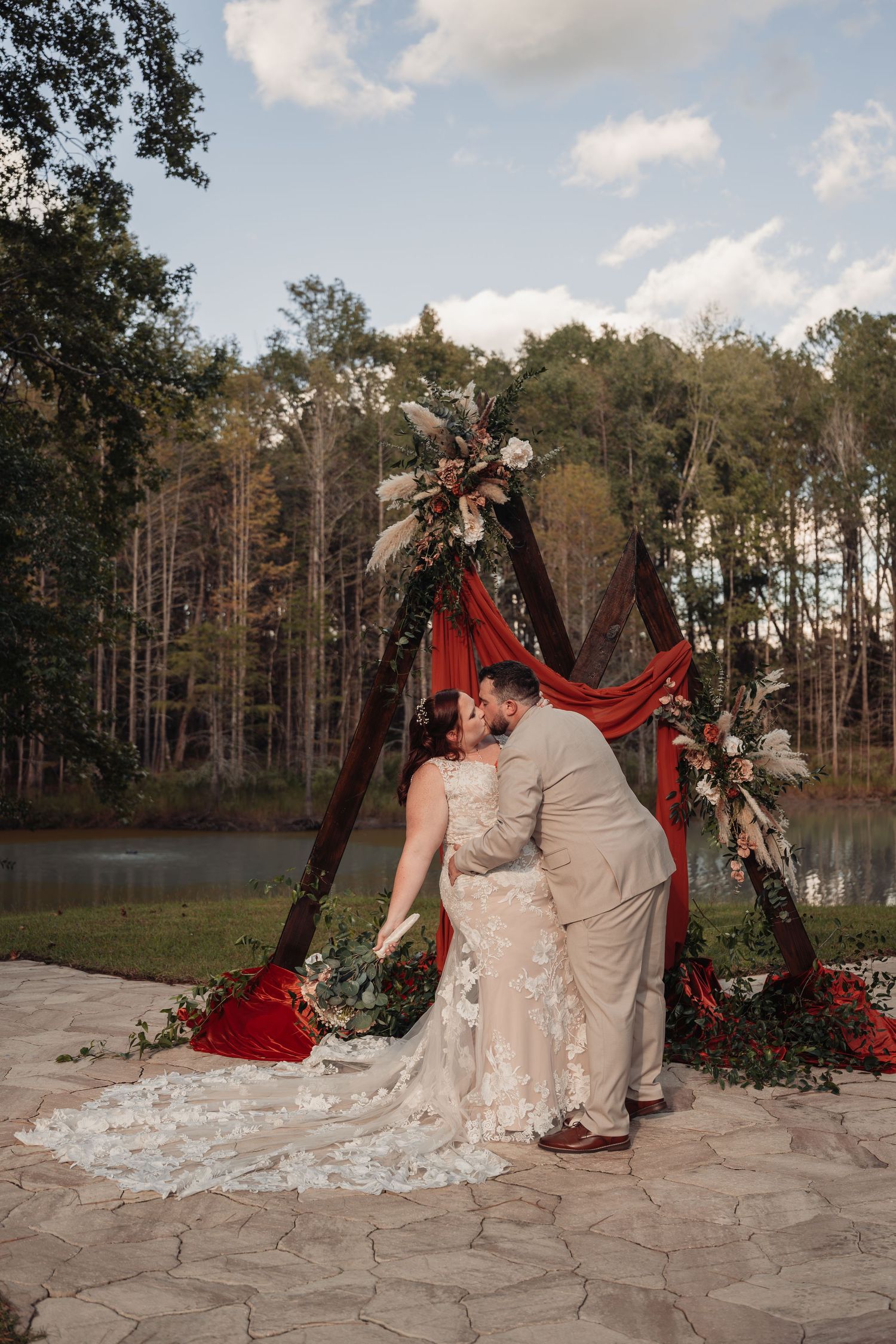 Bride in a lace gown and groom in a tan suit kiss beneath a rustic wooden arch with red drapes and floral accents by a lakeside forest.