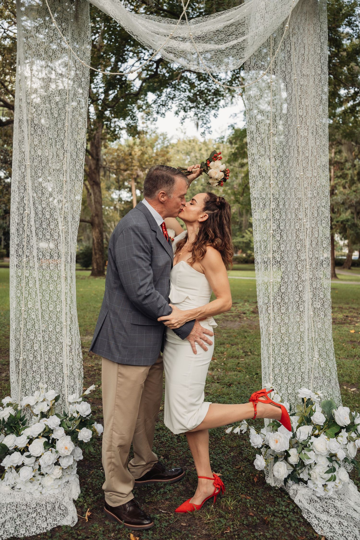 A couple shares their first kiss as newlyweds under a lace arch adorned with white roses and floral draping.