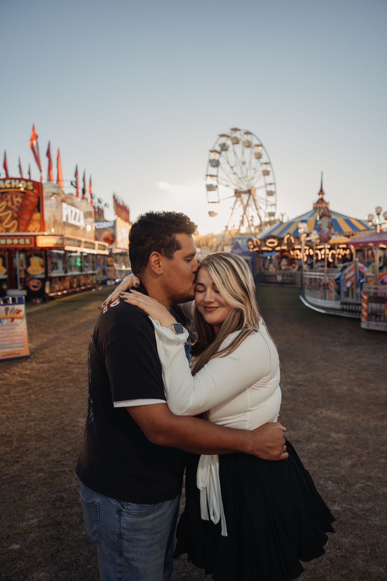 Couple embracing at a sunset carnival as the man kisses the woman's forehead, with a Ferris wheel and festive game booths behind them.